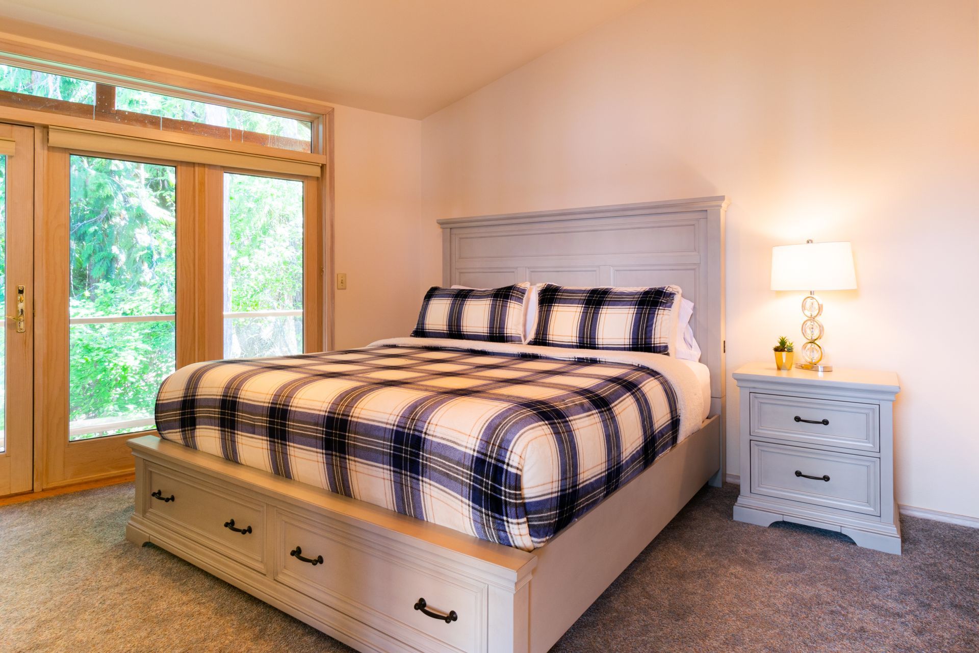 Bedroom with a large bed covered in a blue and white plaid comforter, nightstand with a lamp, and large doors to the outside.