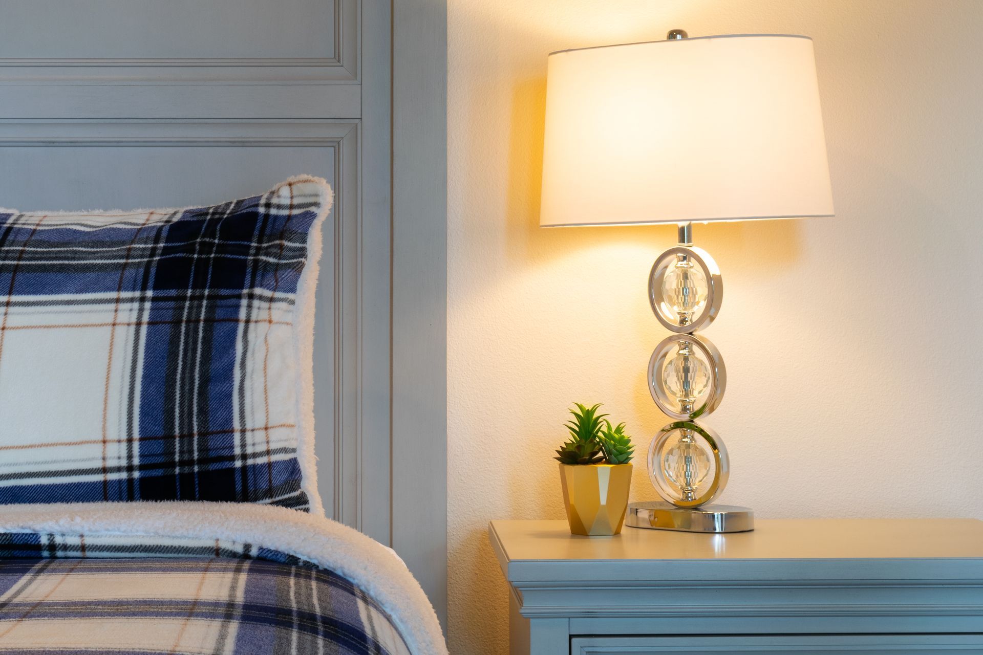 Bedside table with a lit lamp and small plant next to a bed with a blue plaid pillow and comforter.