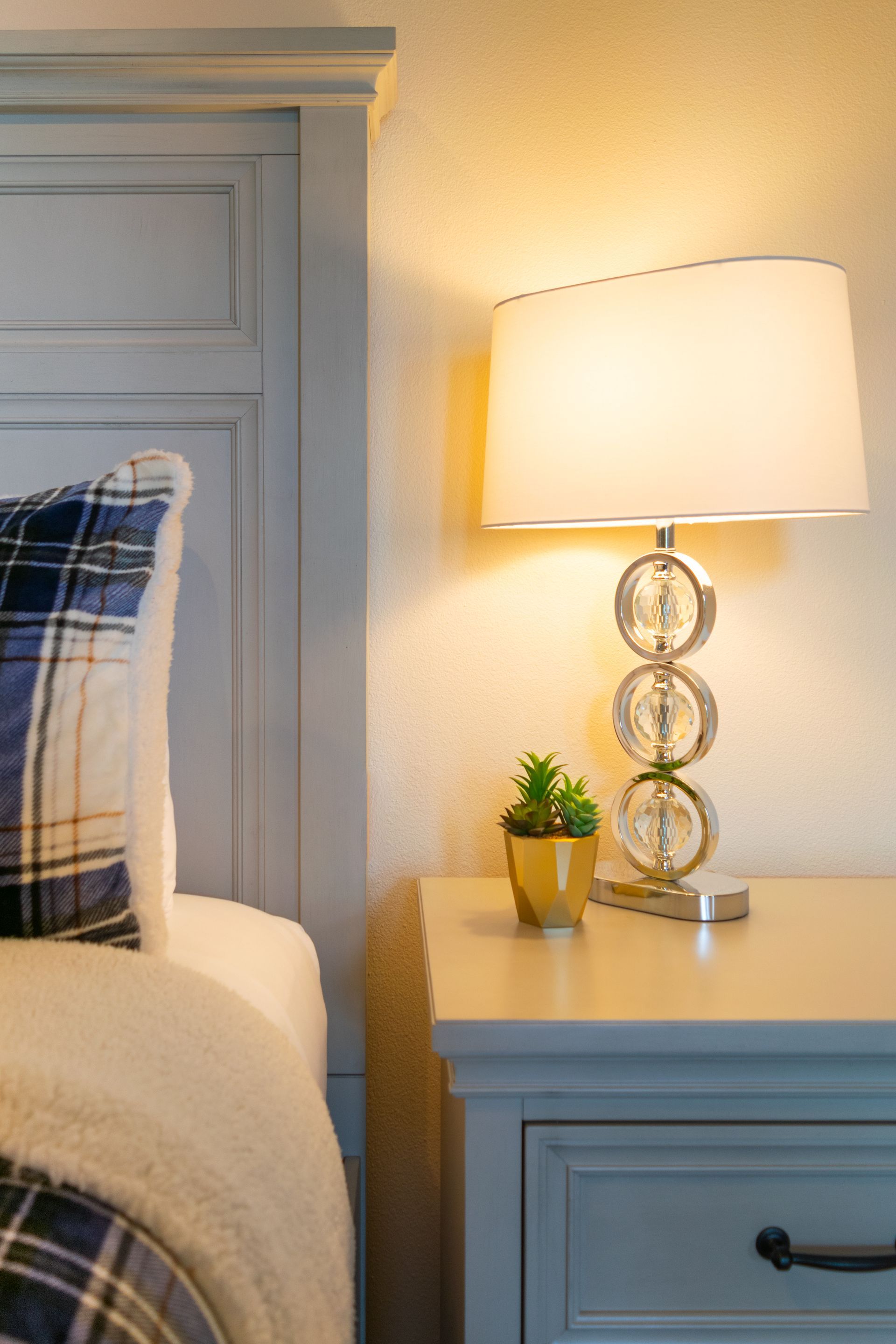 A bedroom nightstand with a lamp, plant, and a plaid pillow next to a bed.