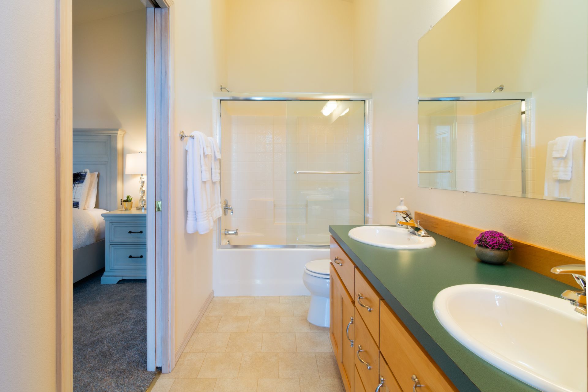 Bathroom with double sinks, toilet, and a bathtub/shower combo. A bedroom is visible through the open door.