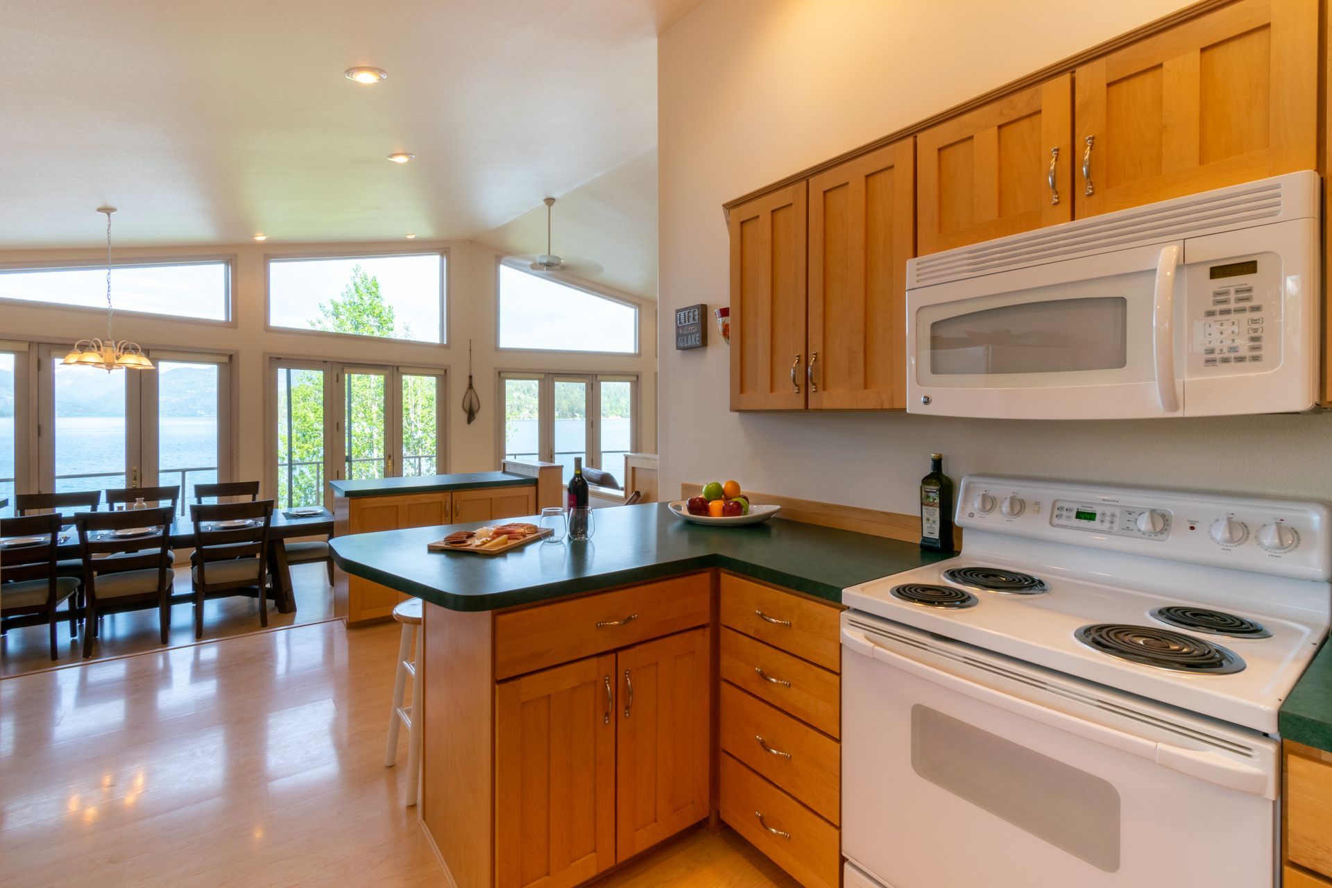 A bright kitchen with wood cabinets, a green countertop, and a white stove. A dining area with a large window is visible.