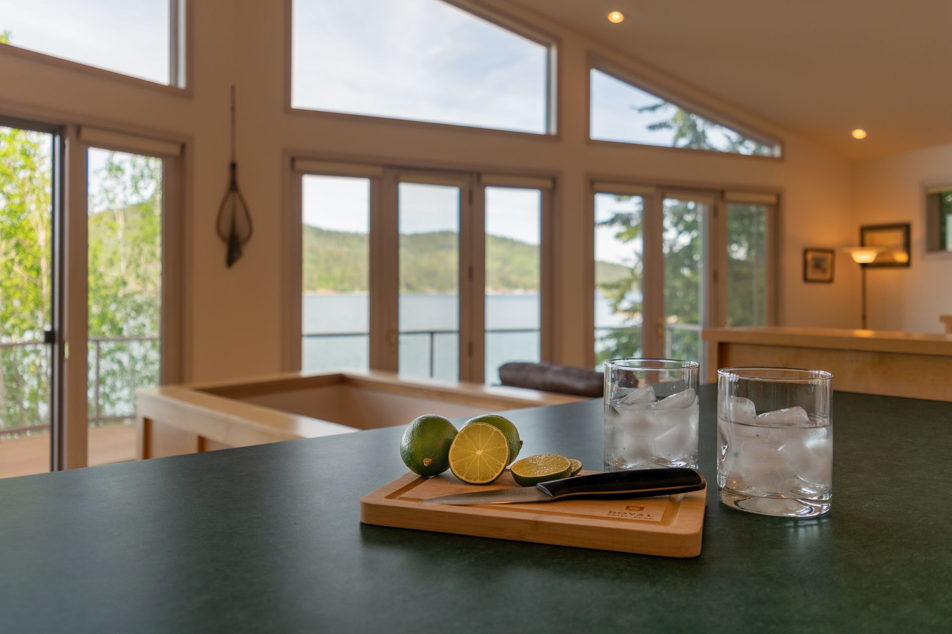 Limes and glasses with ice on a counter in a bright room overlooking a lake. Wooden cutting board and knife are present.