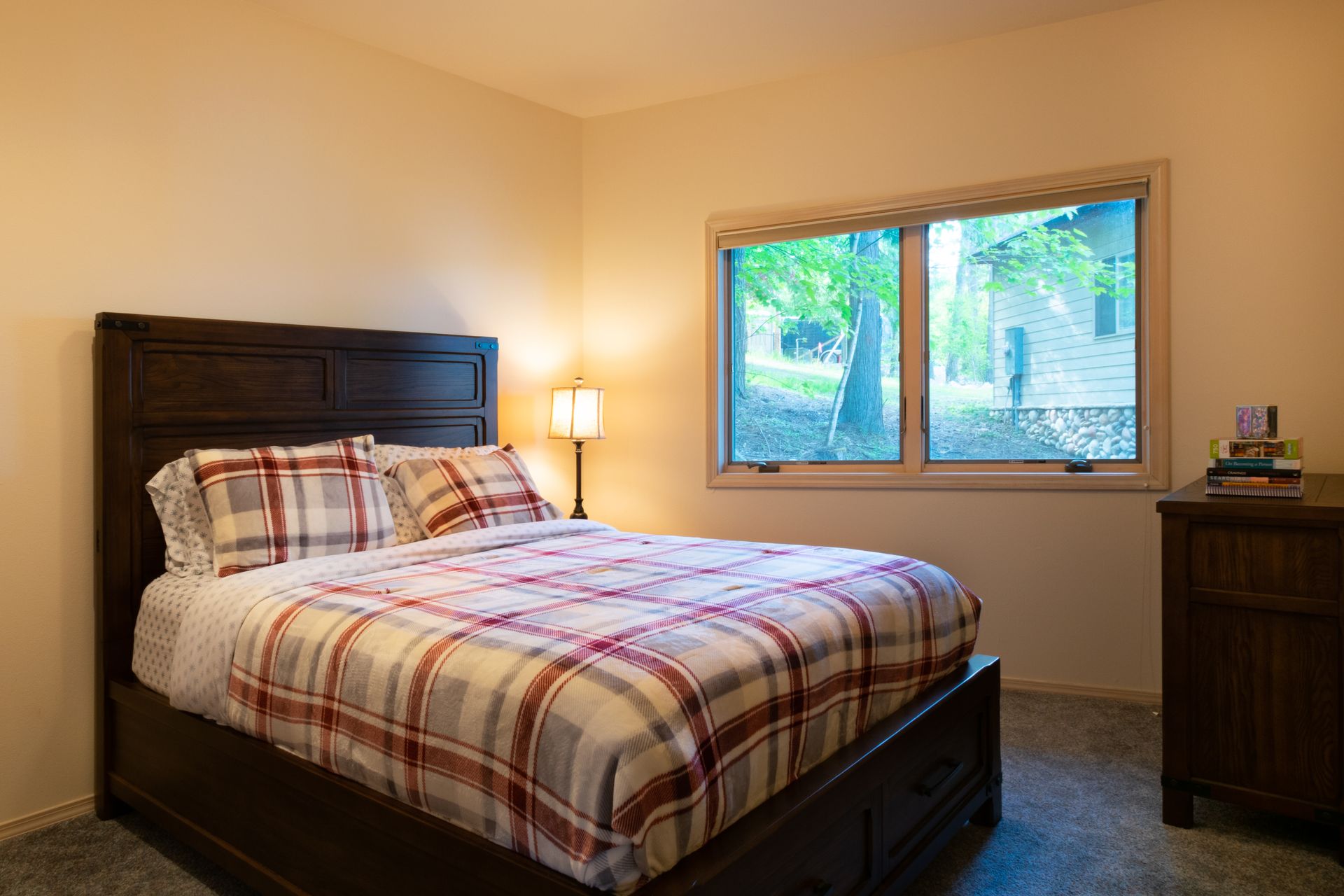 Bedroom with a plaid-covered bed, dark wooden furniture, and a window overlooking trees. A lamp sits on the bedside table.