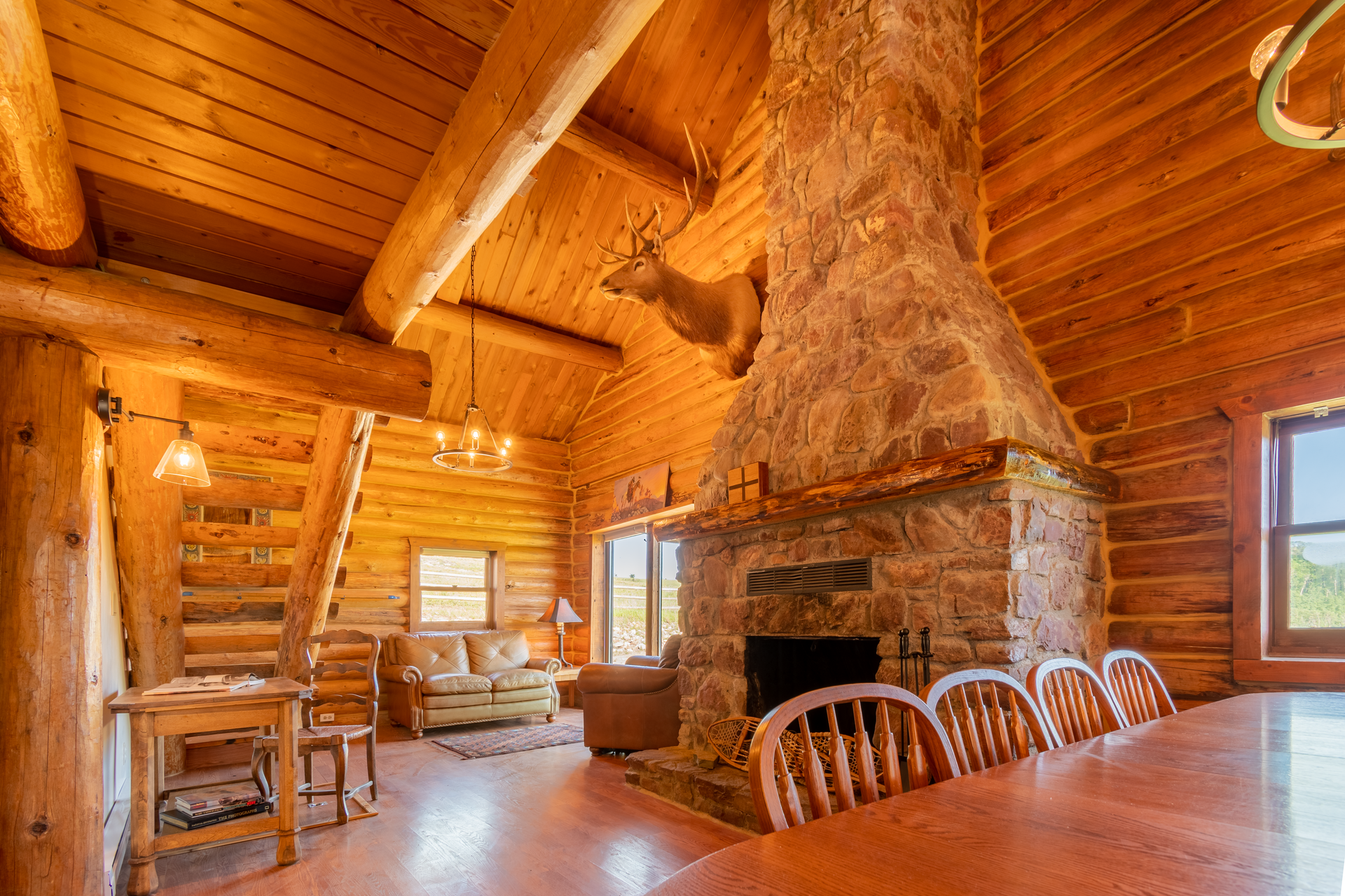 Cozy log cabin interior with a stone fireplace, a dining area, and a taxidermied deer head mounted above the mantel.