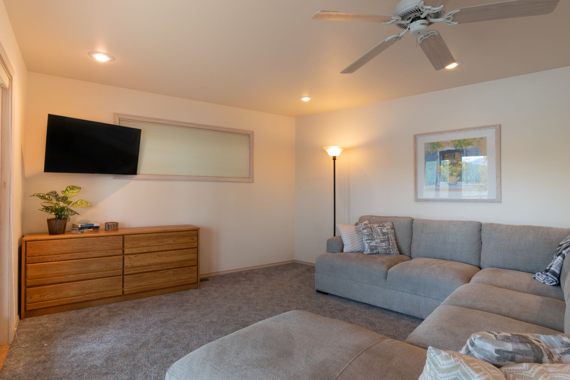 A living room with gray carpet, a gray sectional sofa, and a wooden dresser with a TV above it.