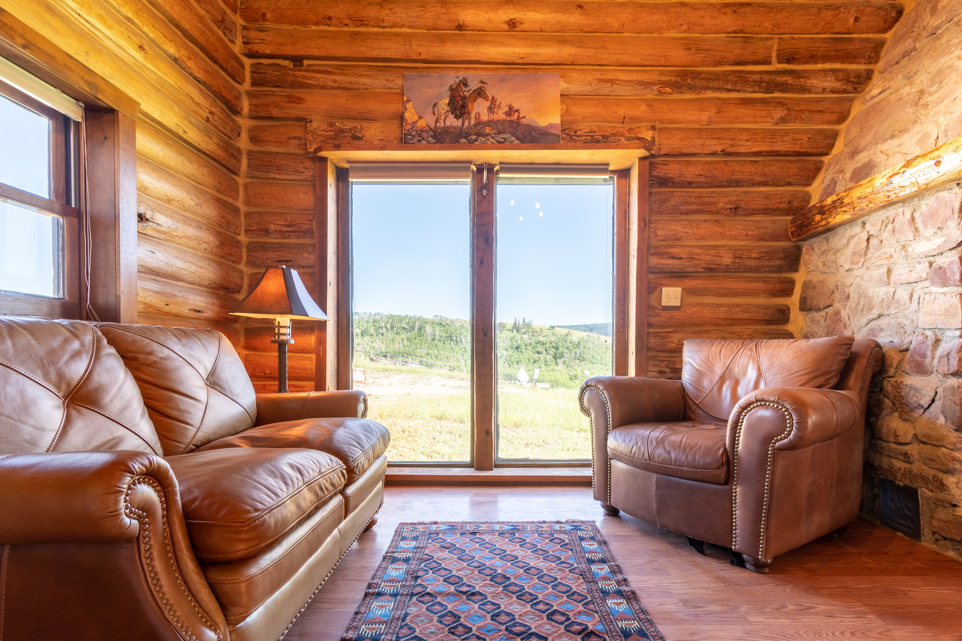 Cozy interior with brown leather sofa and armchair, a large window overlooking a green landscape, and wooden walls.
