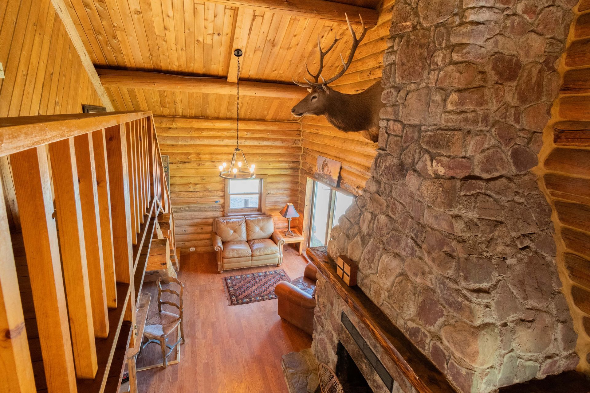 Interior of a log cabin living room with a stone fireplace and a mounted elk head. A loft overlooks the space.
