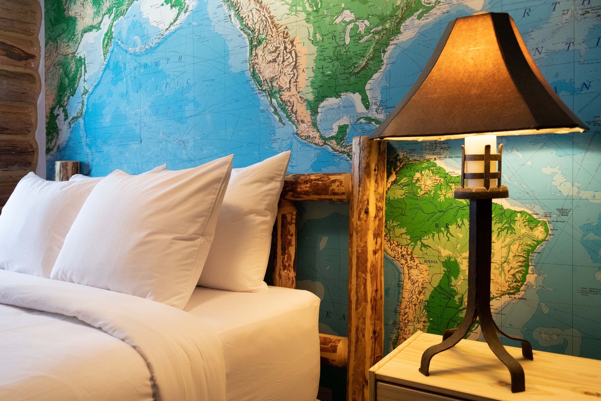A bedroom with a world map mural headboard, white bedding, and a lit lamp on a nightstand.