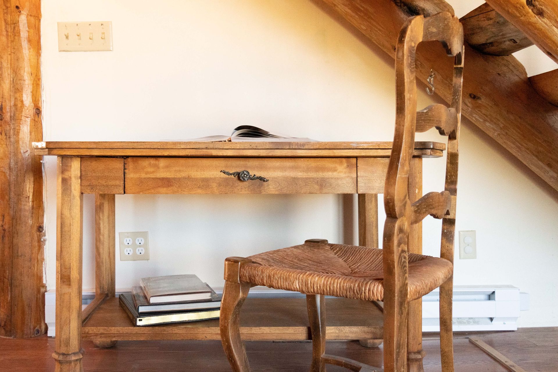 Wooden desk and chair in a cozy room with a natural wood staircase and a stack of books underneath the desk.