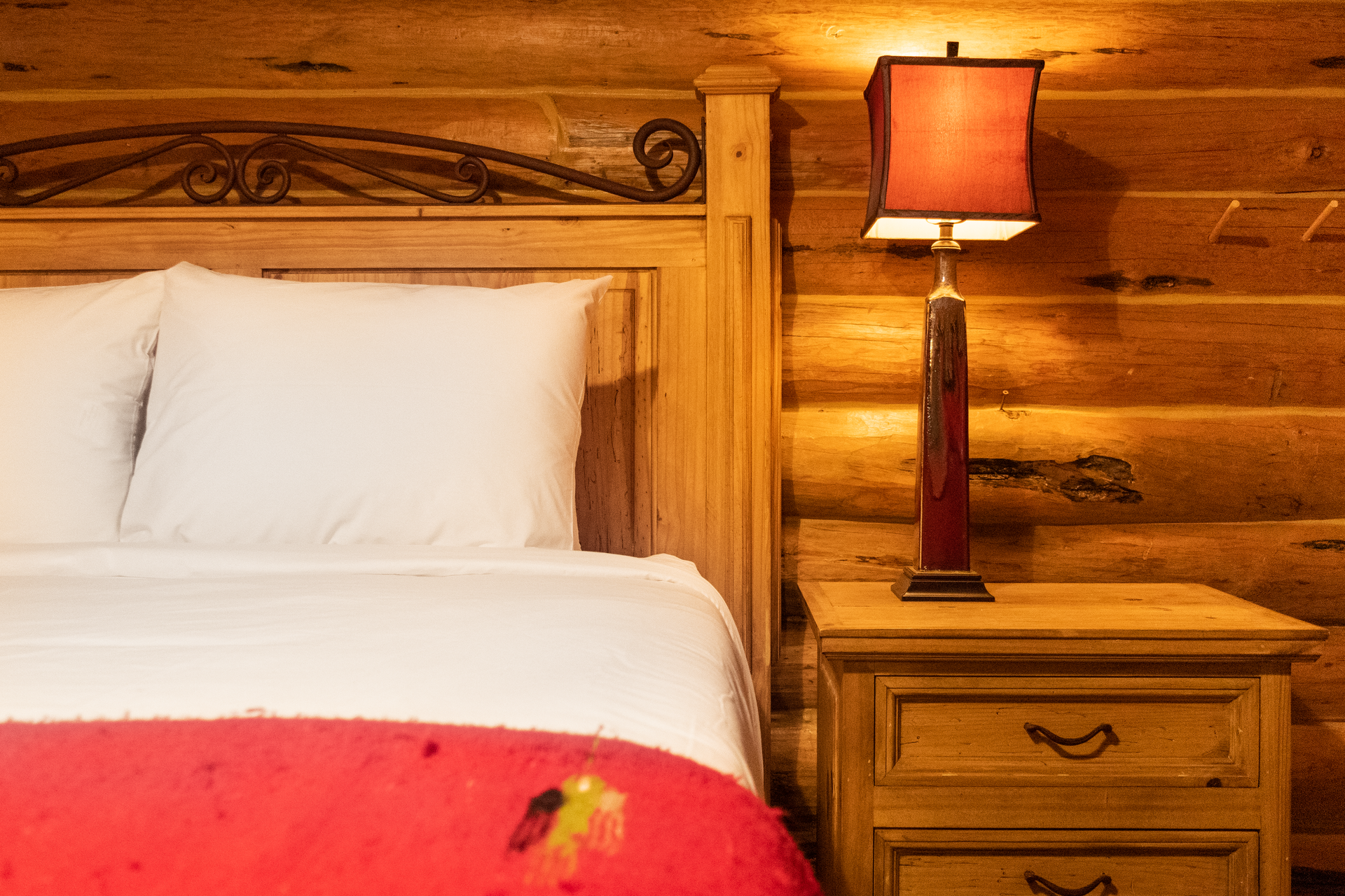 Wooden bedroom with a bed, nightstand, and lamp. The room has log walls and the bed has white linens and a red blanket.