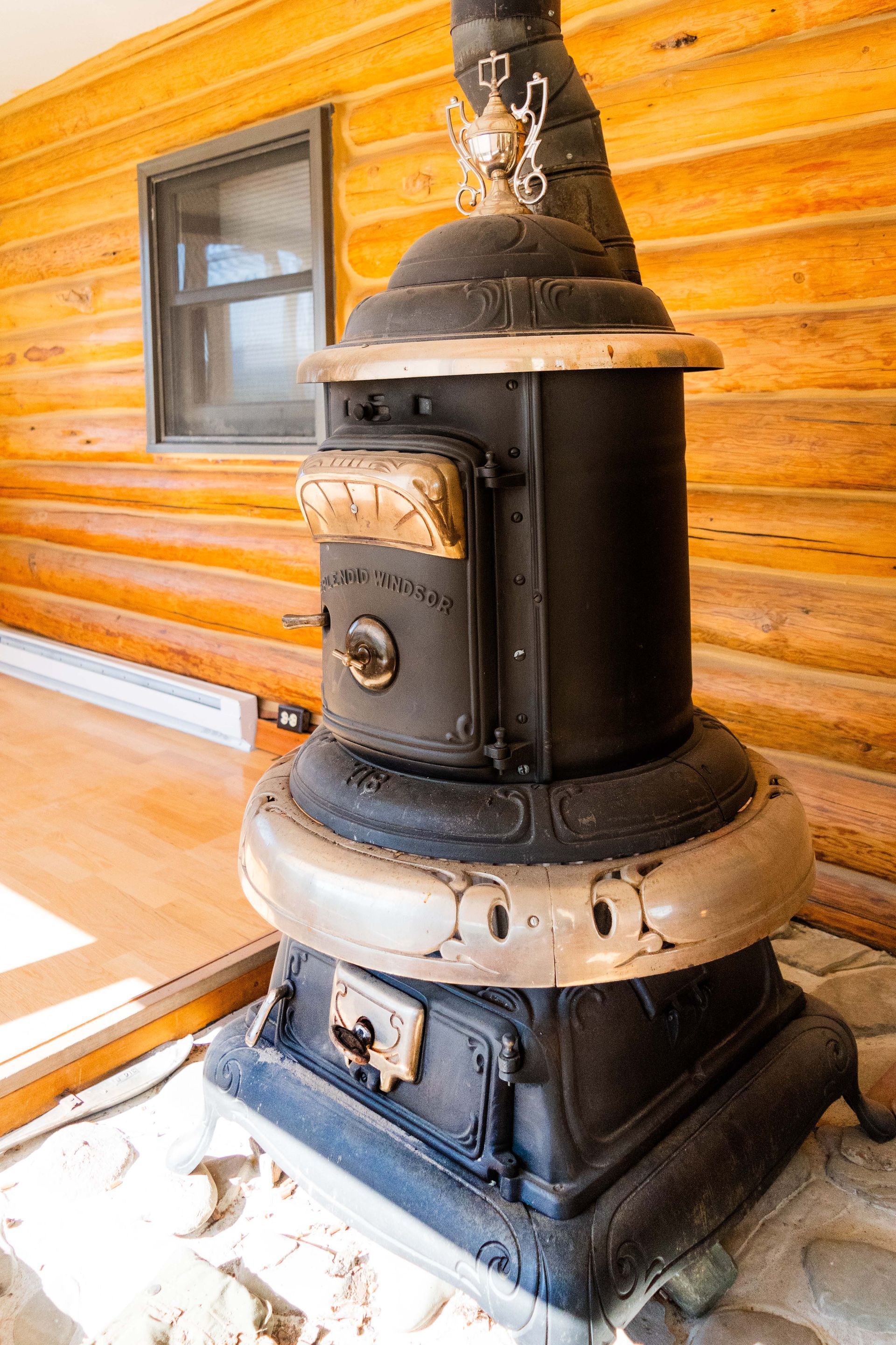 Black antique wood-burning stove with a silver trim, against a wooden cabin wall next to a small window.