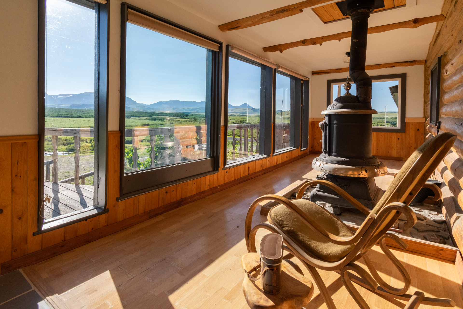 A sunroom with large windows overlooking a landscape. A wooden rocking chair sits next to an old stove.