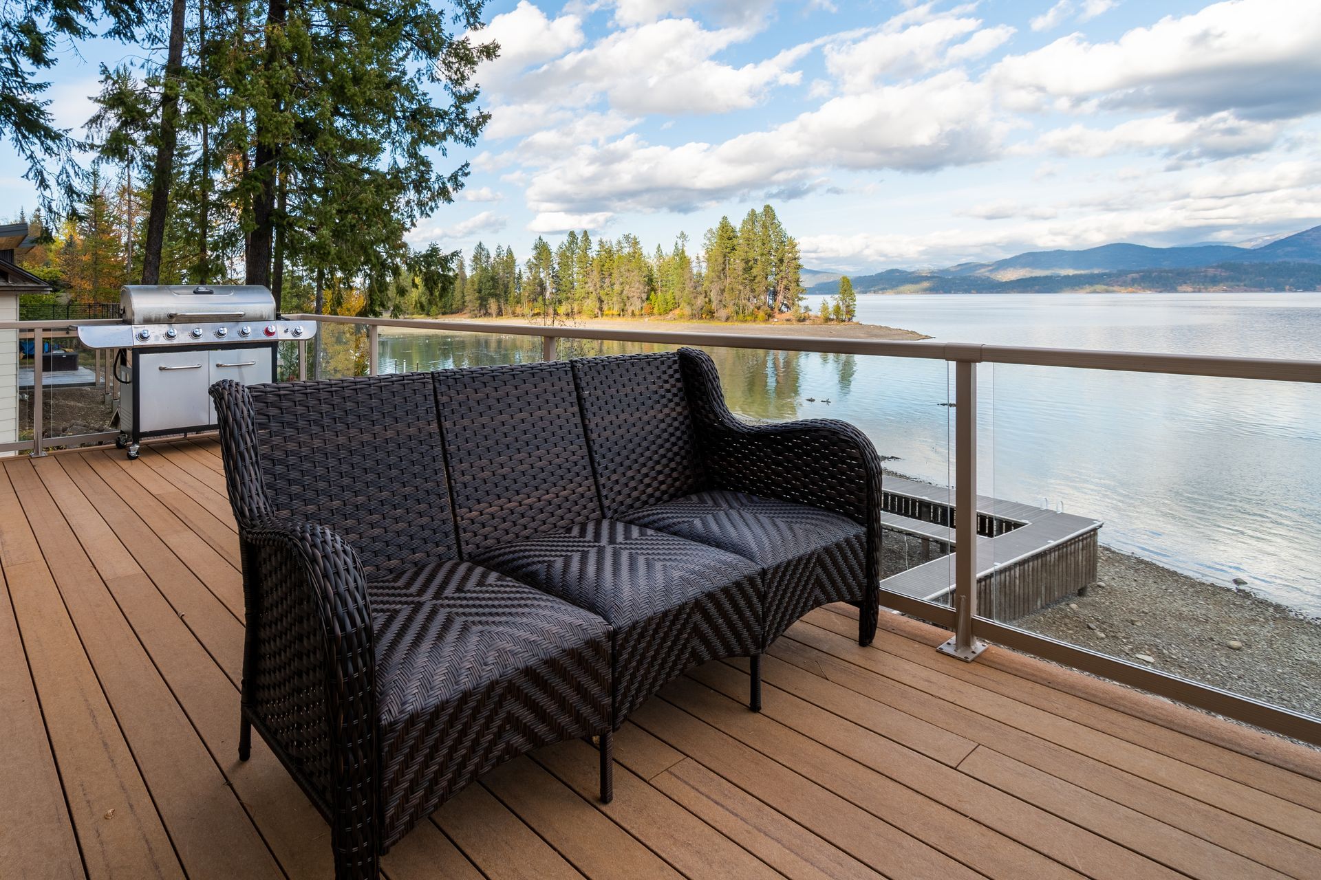 Wicker couch on a wooden deck overlooking a lake, with a grill, glass railing, and trees in the background.