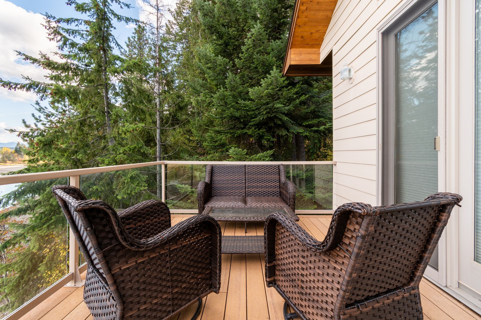 A deck with wicker furniture overlooking a green forest and water. The house has white siding and a glass railing.