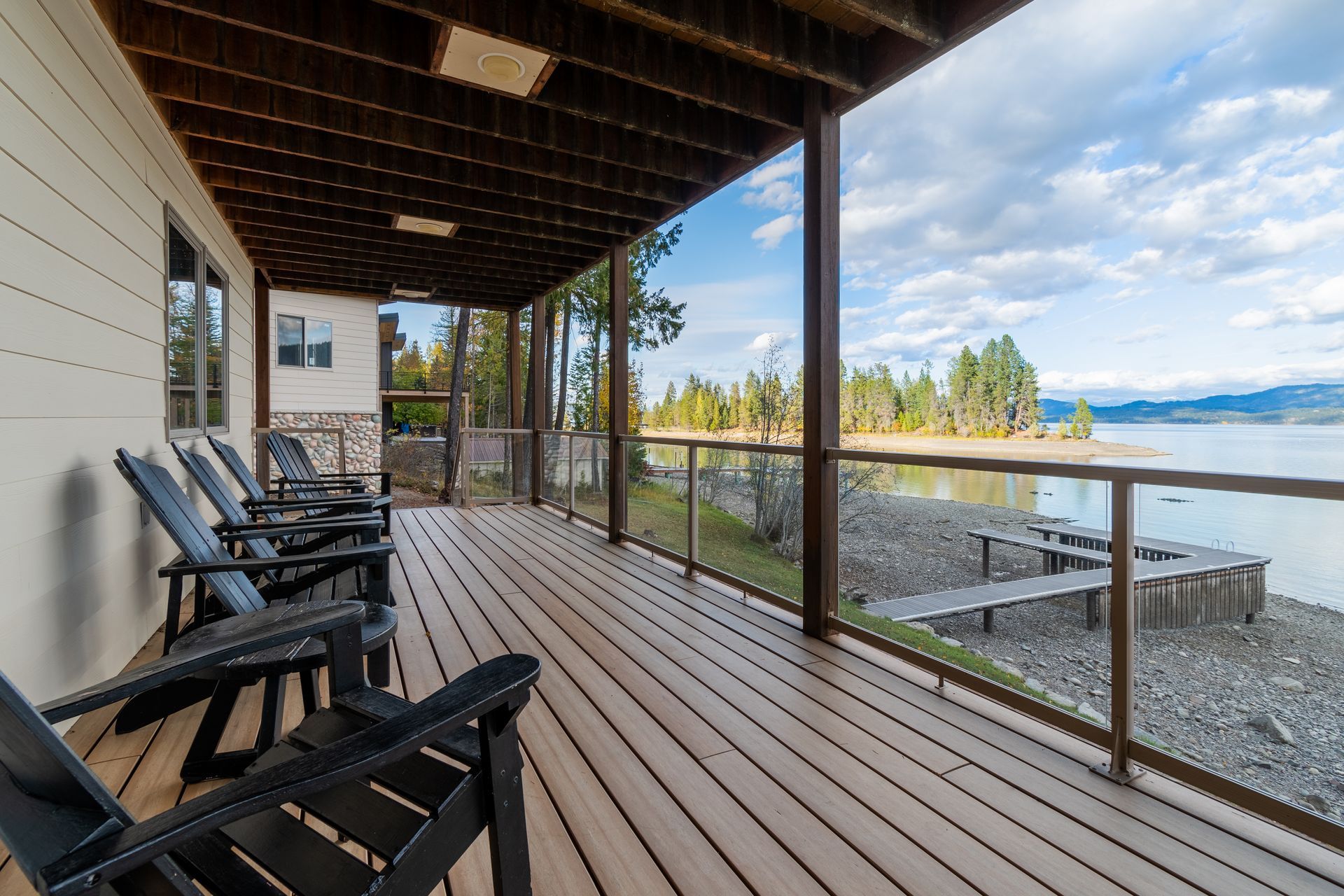 A wooden deck with black chairs overlooks a lake with a wooden dock and a cloudy sky.