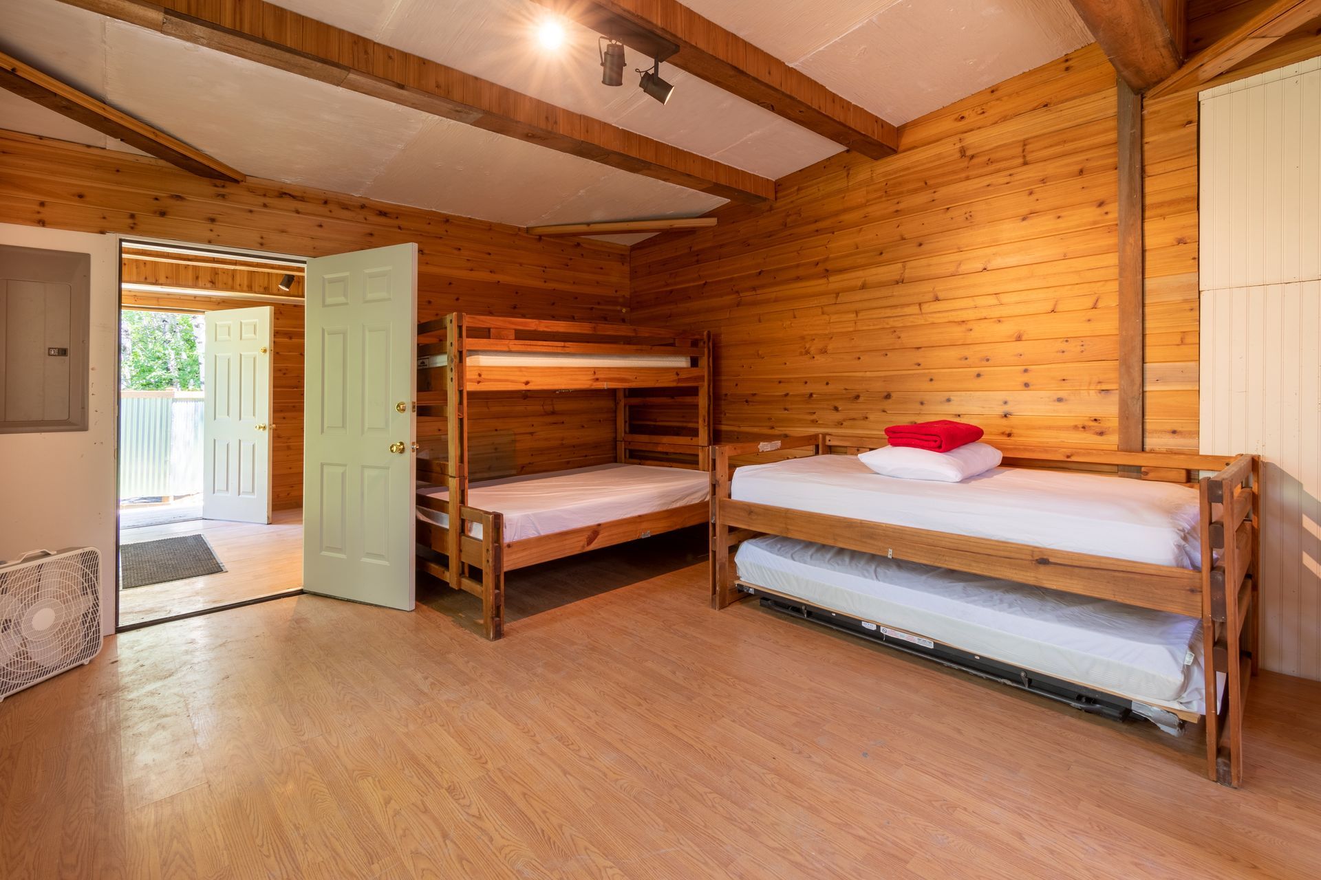 A rustic cabin bedroom with wooden walls and floor. There are bunk beds and a bed with a trundle, along with an open doorway.