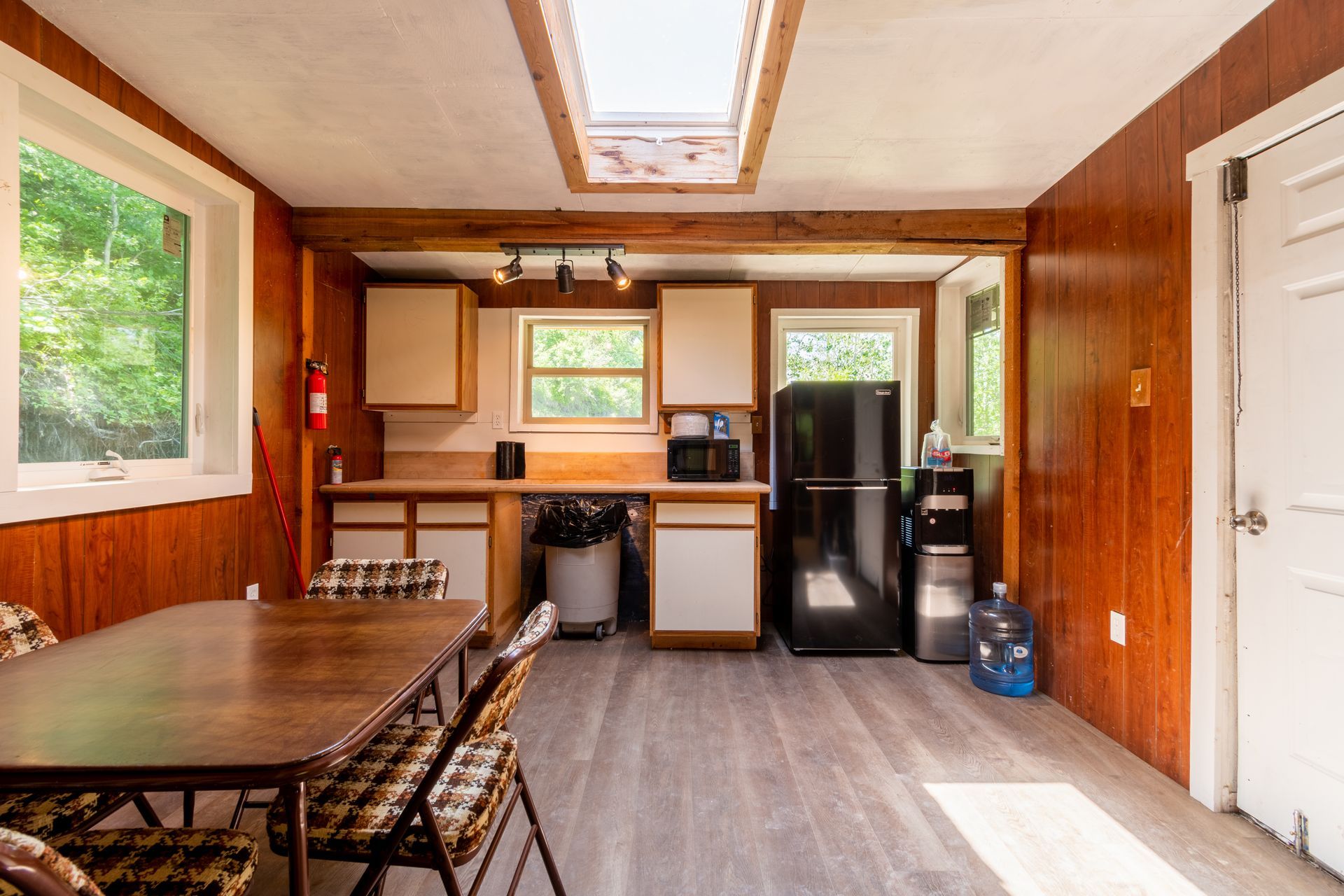 A rustic kitchen with wood paneling, white cabinets, and a dining table. A black refrigerator stands by a white door.