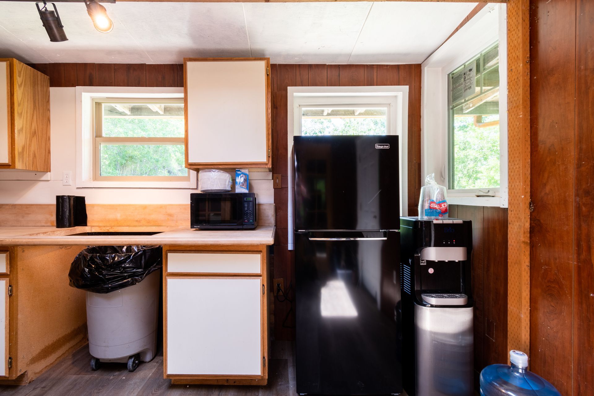 A small kitchen with wood paneling, white cabinets, a black refrigerator, and two windows.