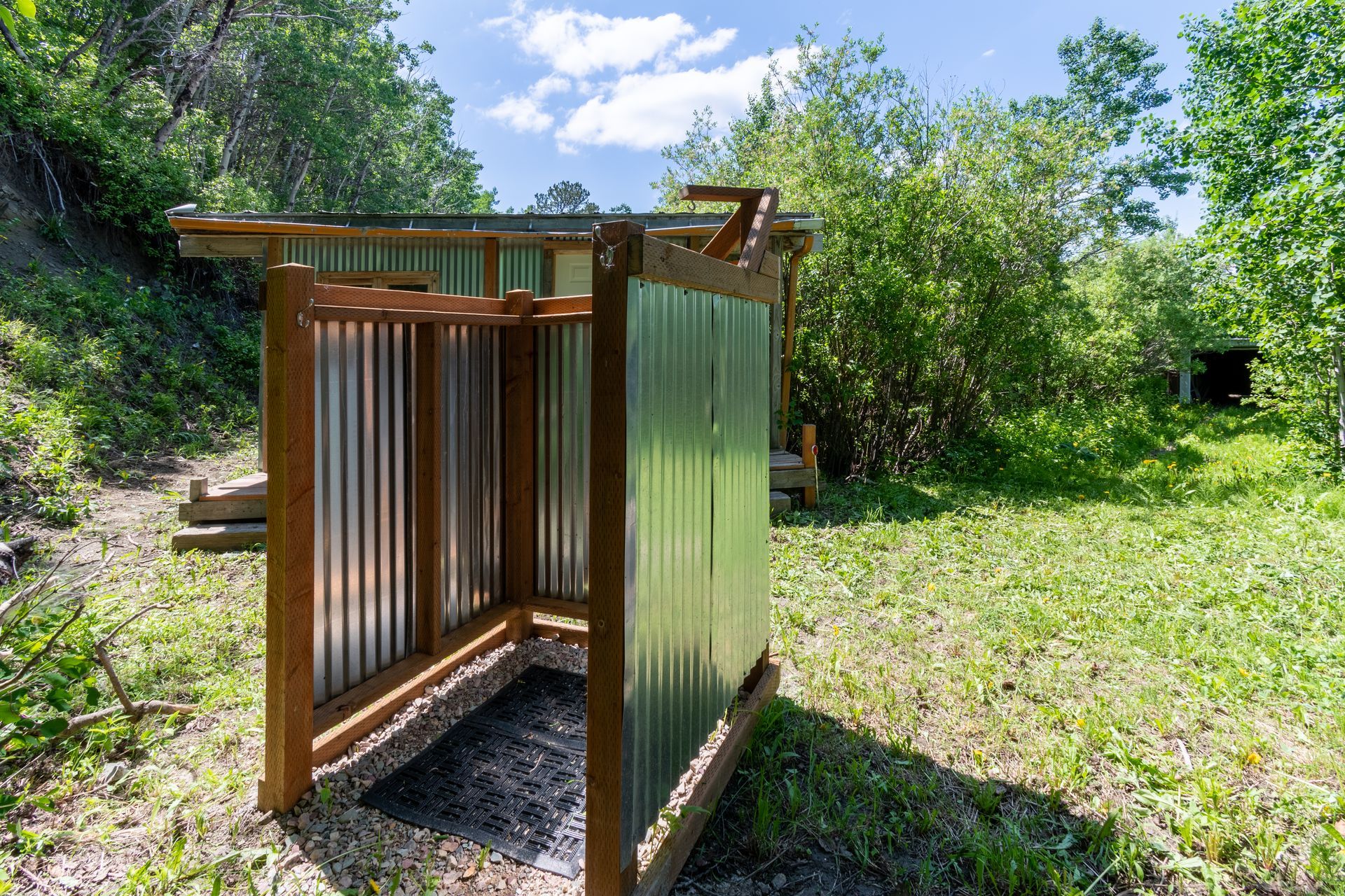 Outdoor shower stall with corrugated metal walls and wooden frame, set in a grassy clearing surrounded by trees.
