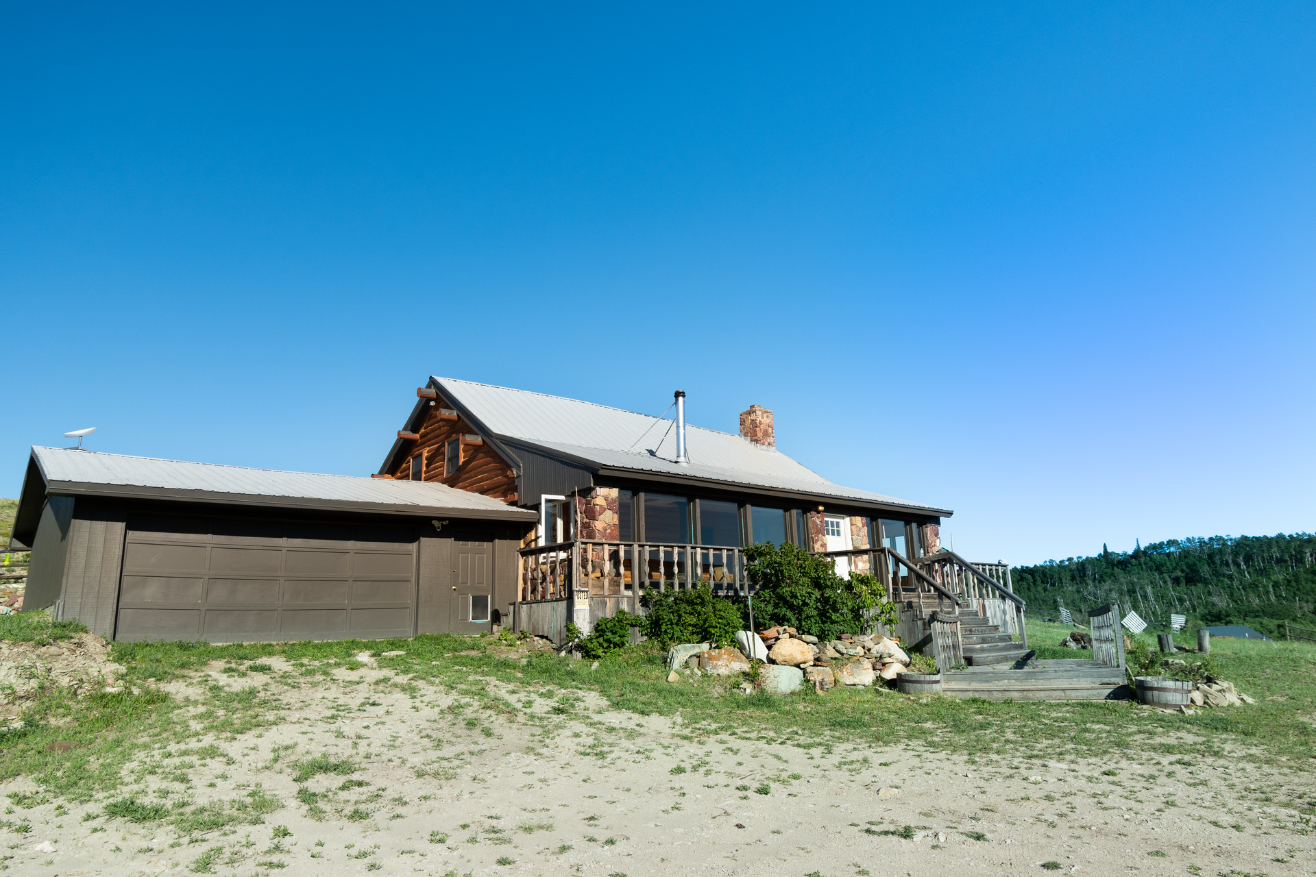 A rustic, wooden house with a garage sits on a grassy hill under a clear blue sky.