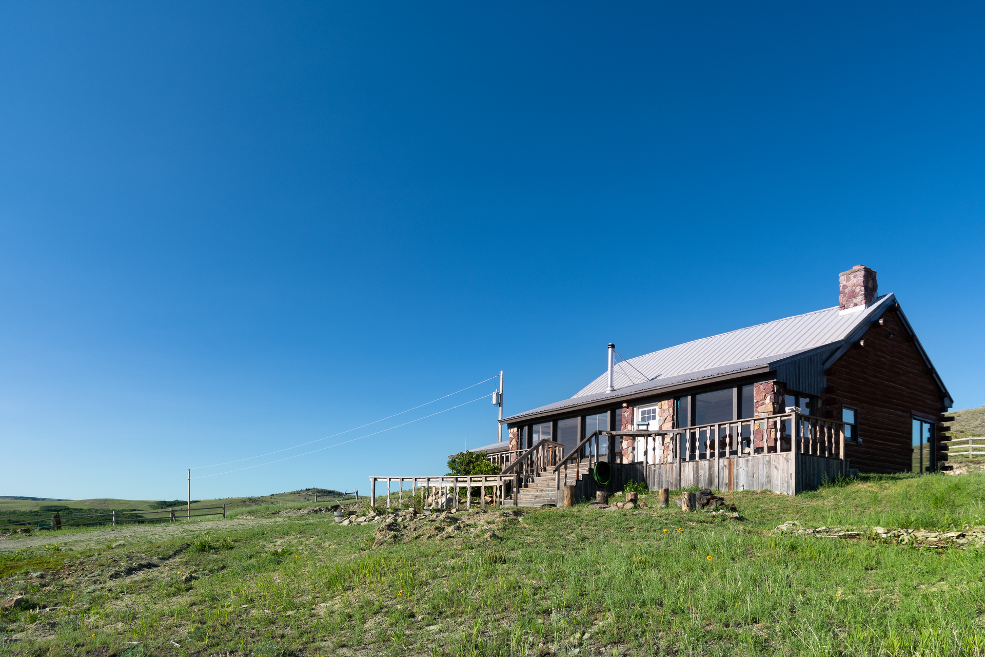 A rustic wooden cabin with a metal roof sits on a grassy hillside under a clear blue sky.