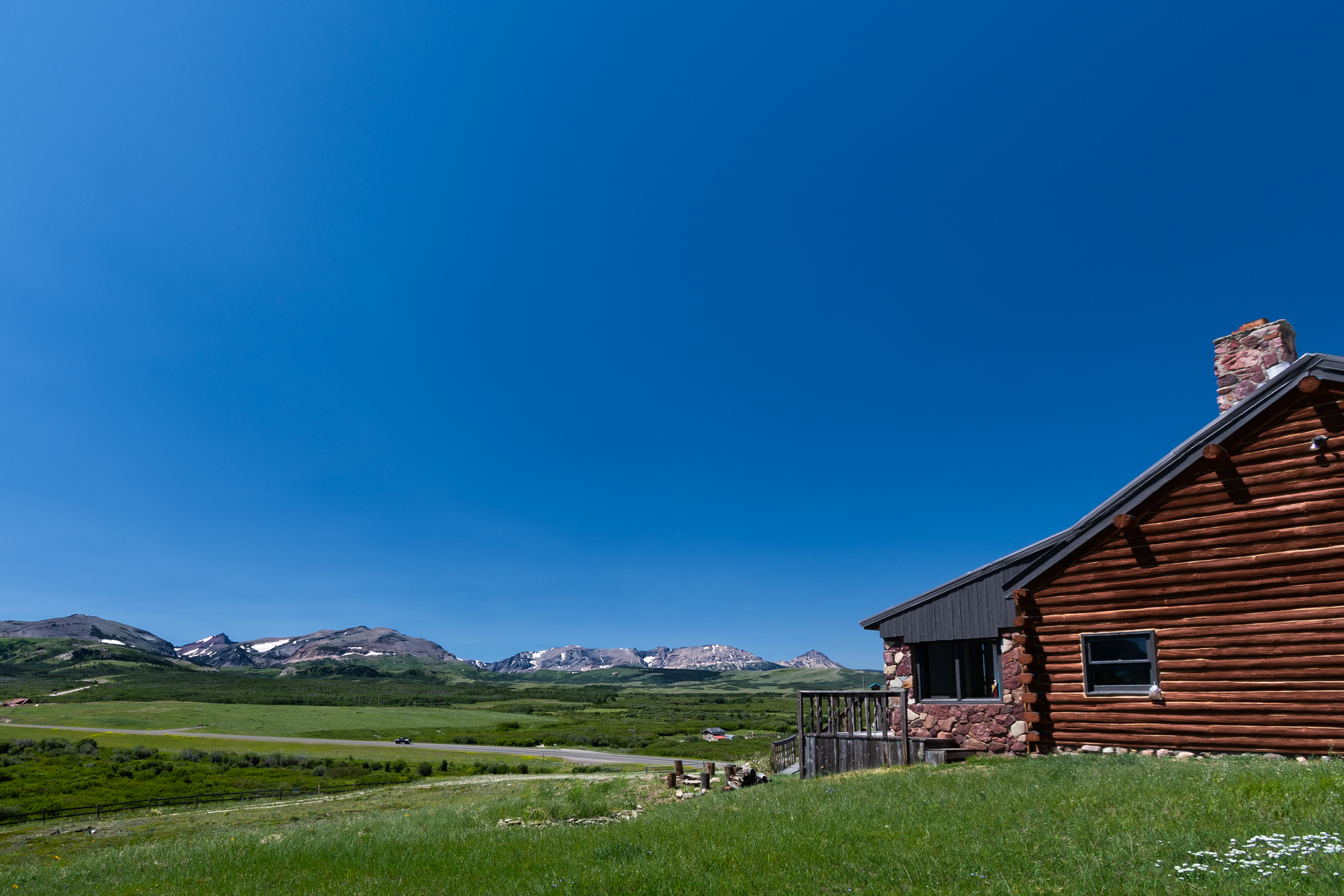 Wooden cabin on a grassy hill with a bright blue sky and distant mountain range in the background.