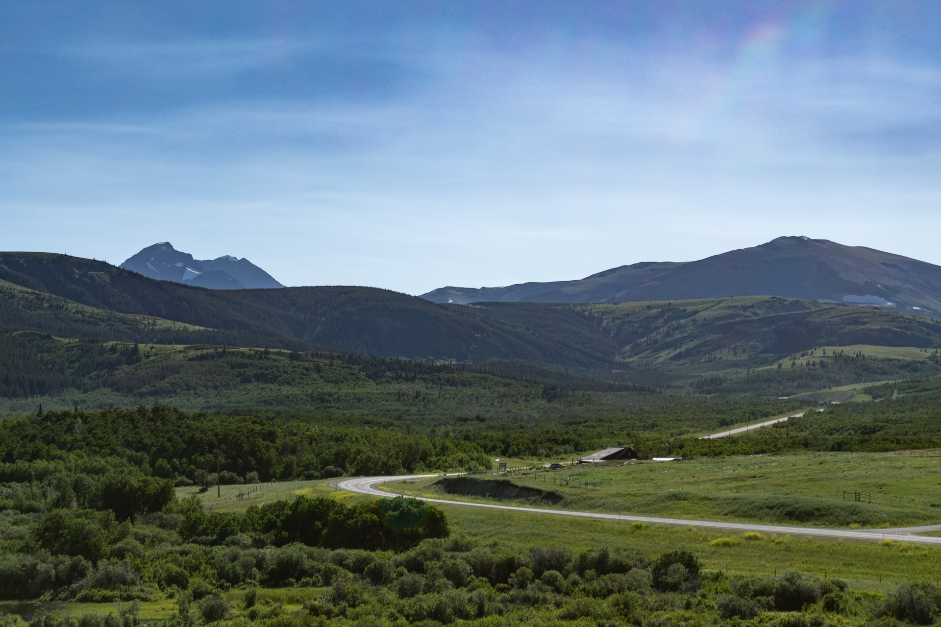 A winding road through a green valley surrounded by mountains under a bright blue sky.