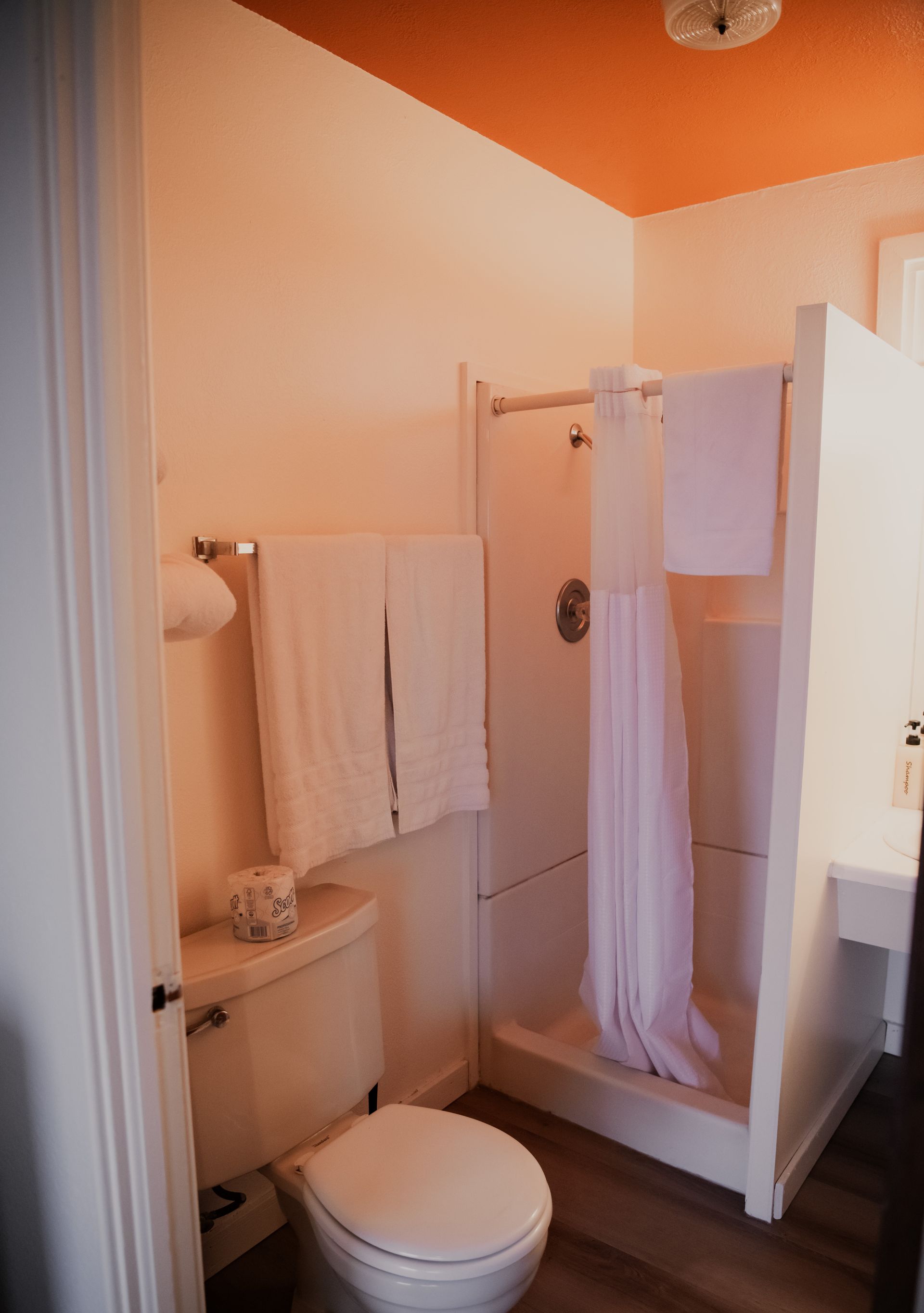 Bathroom with white toilet, shower, and towels; orange ceiling.