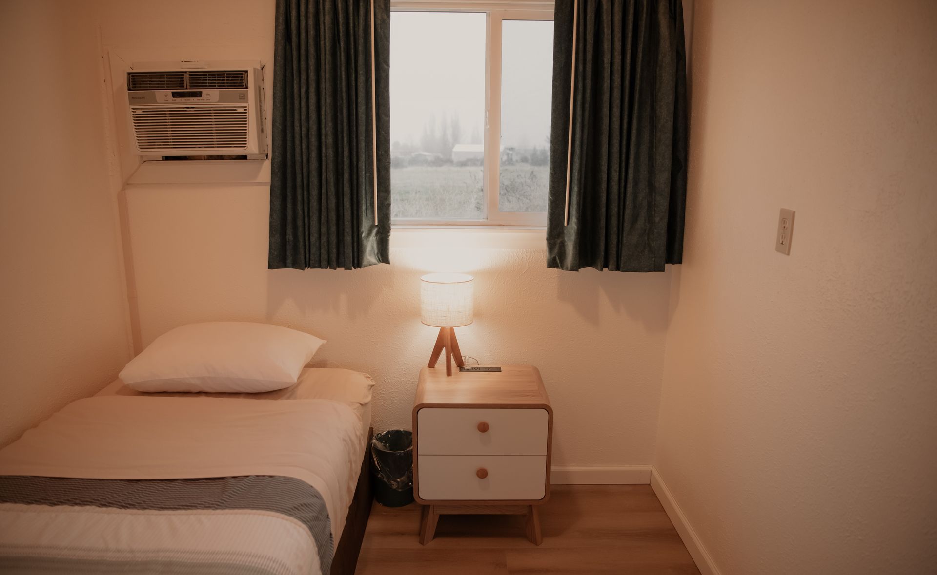 A small, simple bedroom with a twin bed, nightstand, and window with curtains.