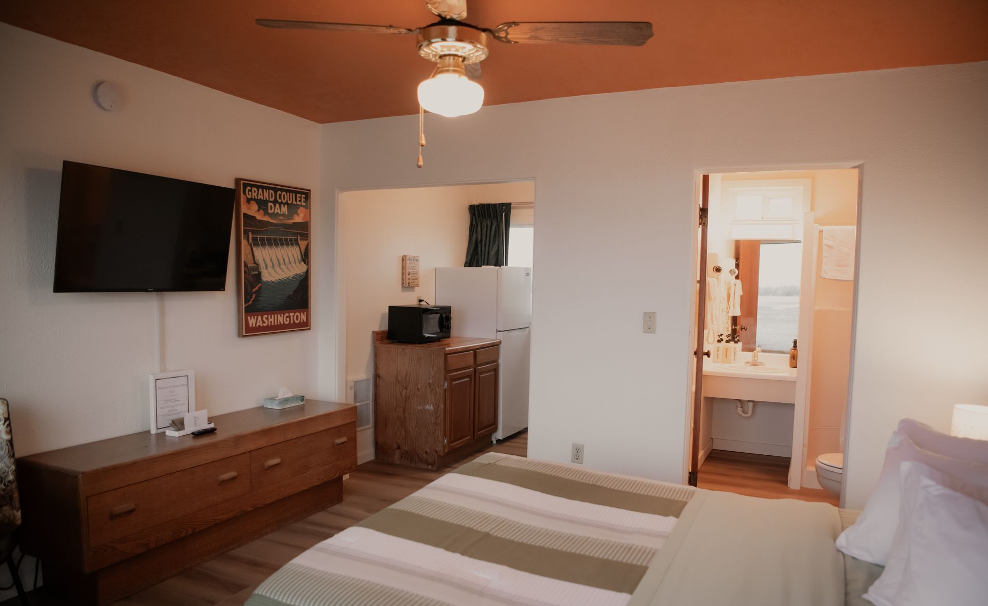 A motel room with a bed, dresser, and a small table. The room has a neutral color palette with green accents.