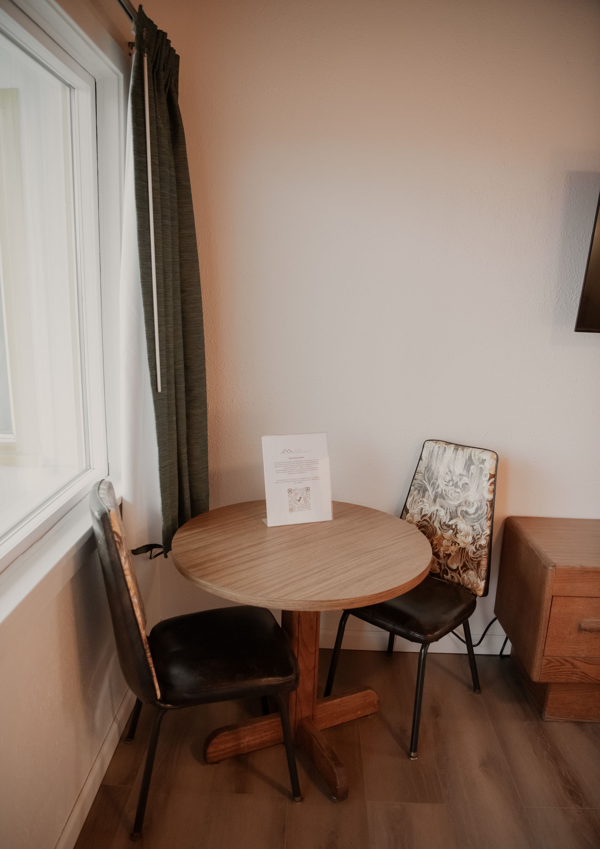Round table with two chairs near a window. A sign sits on the table; a dresser is to the right.