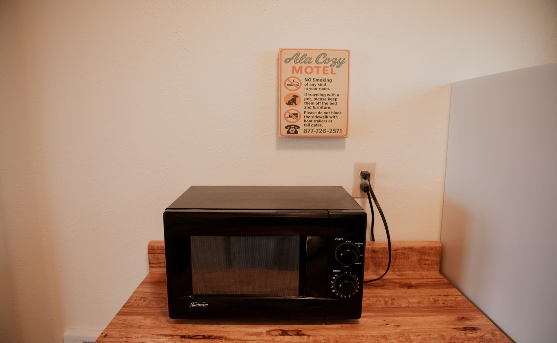 Black microwave on a wooden counter with a sign and electrical outlet on a white wall.