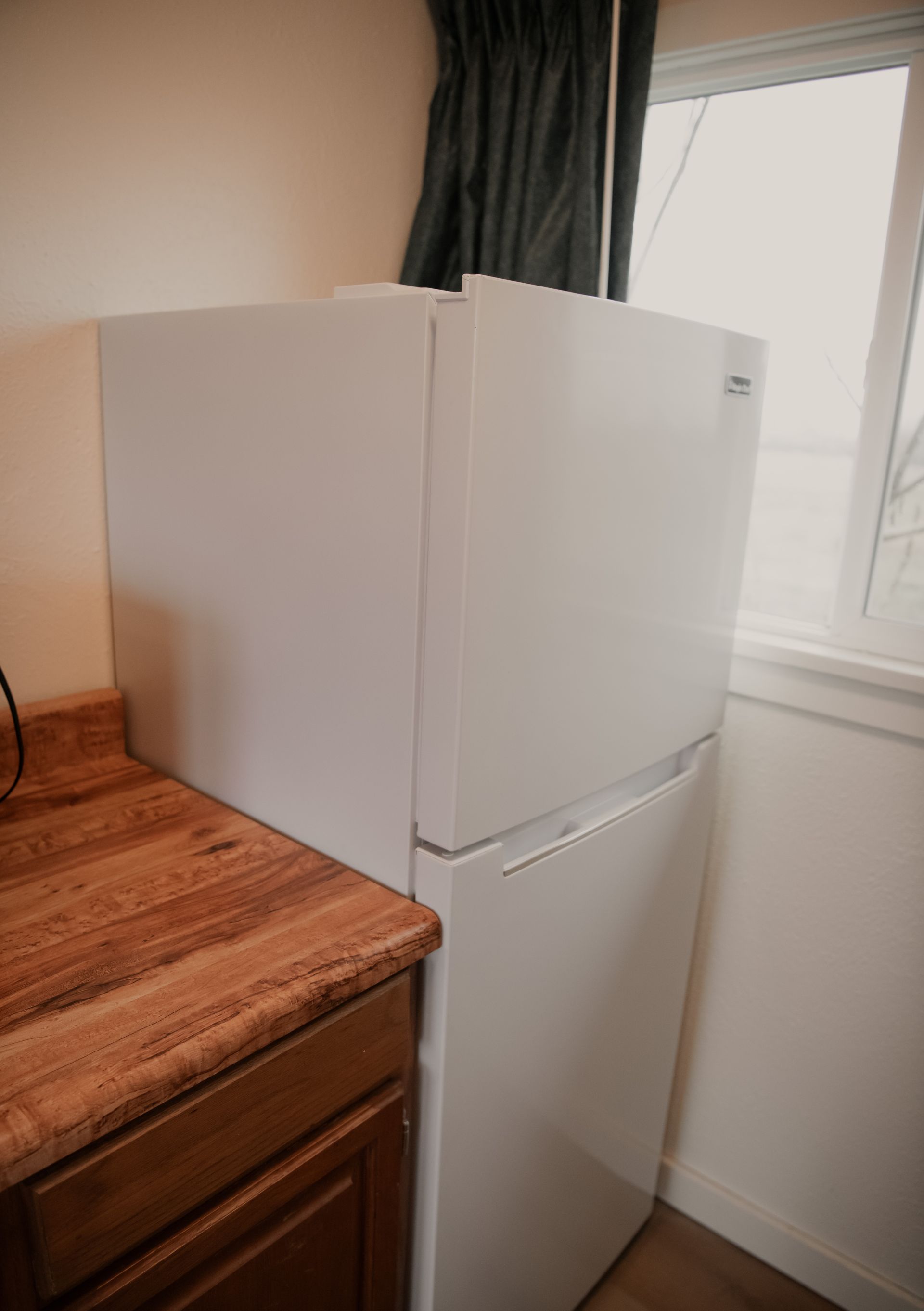 White refrigerator next to a wooden countertop and window with a dark curtain.