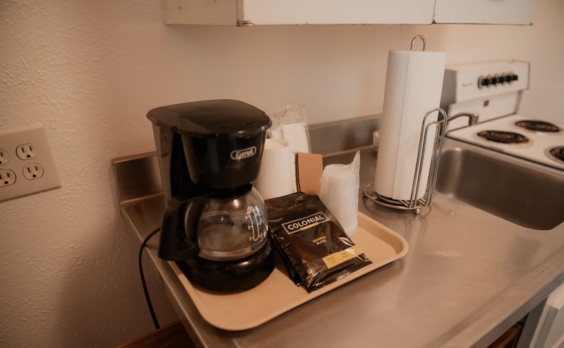 Coffee maker and supplies on a tray next to a kitchen sink and stovetop.