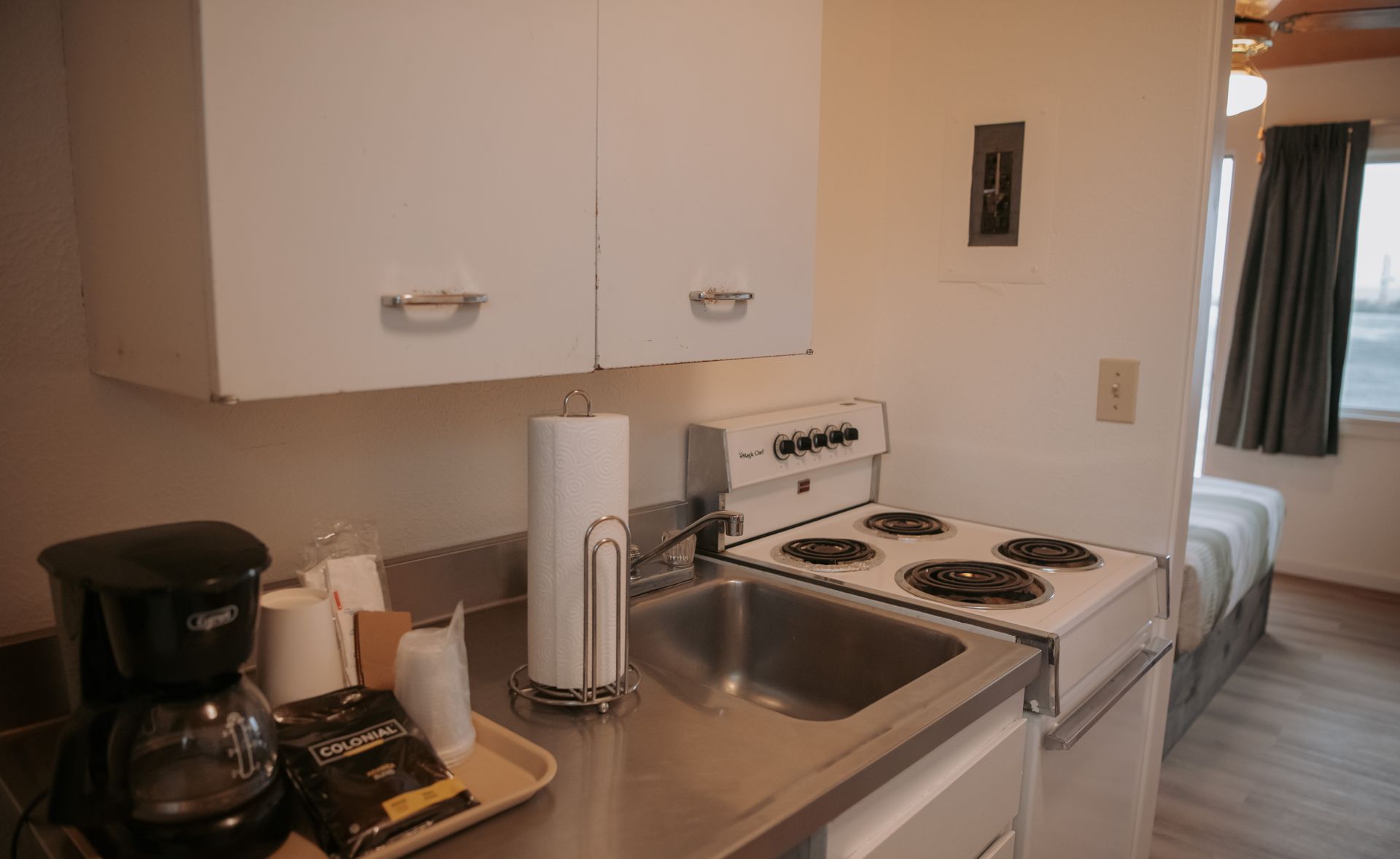 Small kitchen with a stovetop, sink, cabinets, and a coffee maker. A bed is visible in the background.