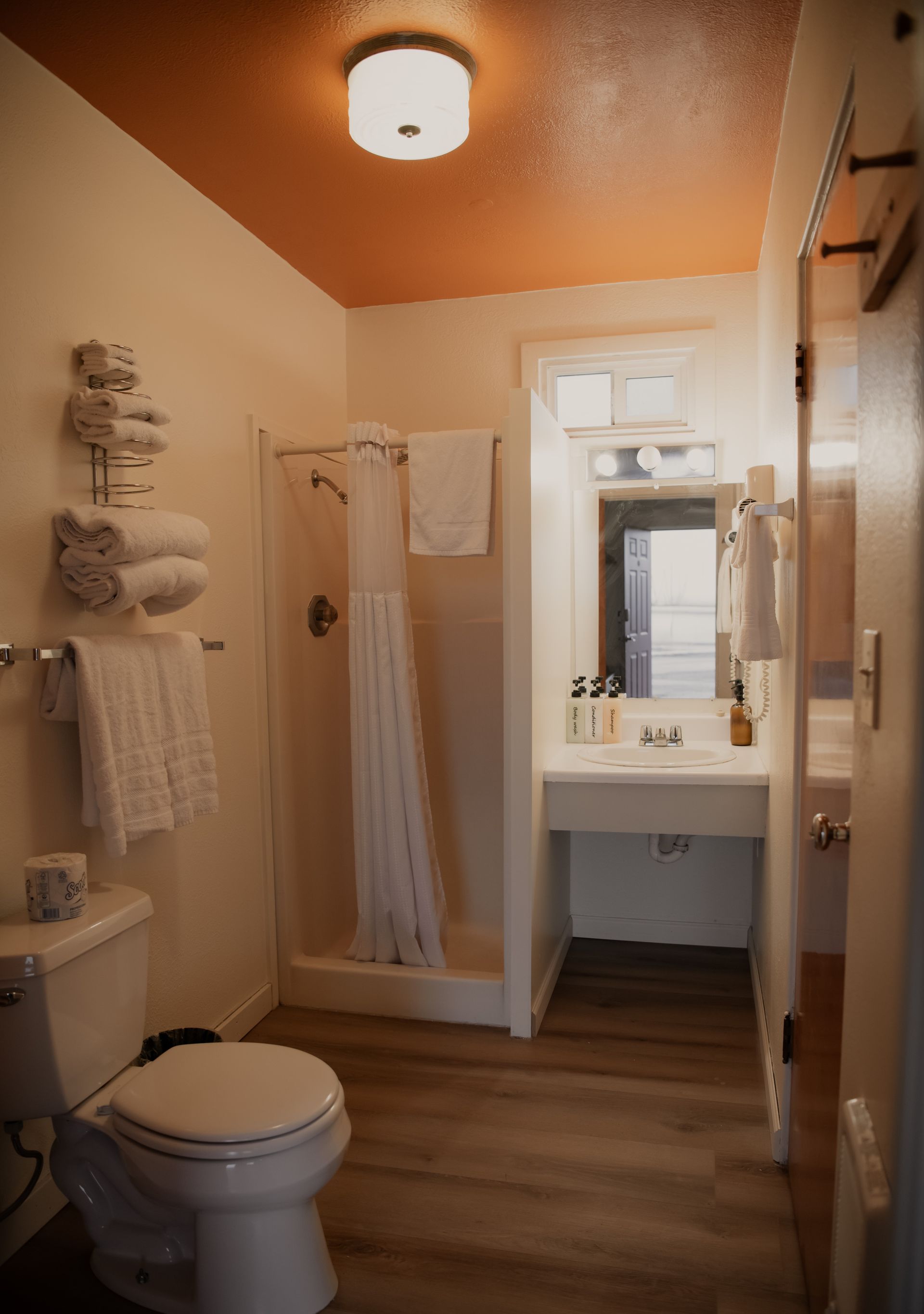 Small bathroom with a toilet, shower, and sink; orange ceiling and light wood-look flooring.