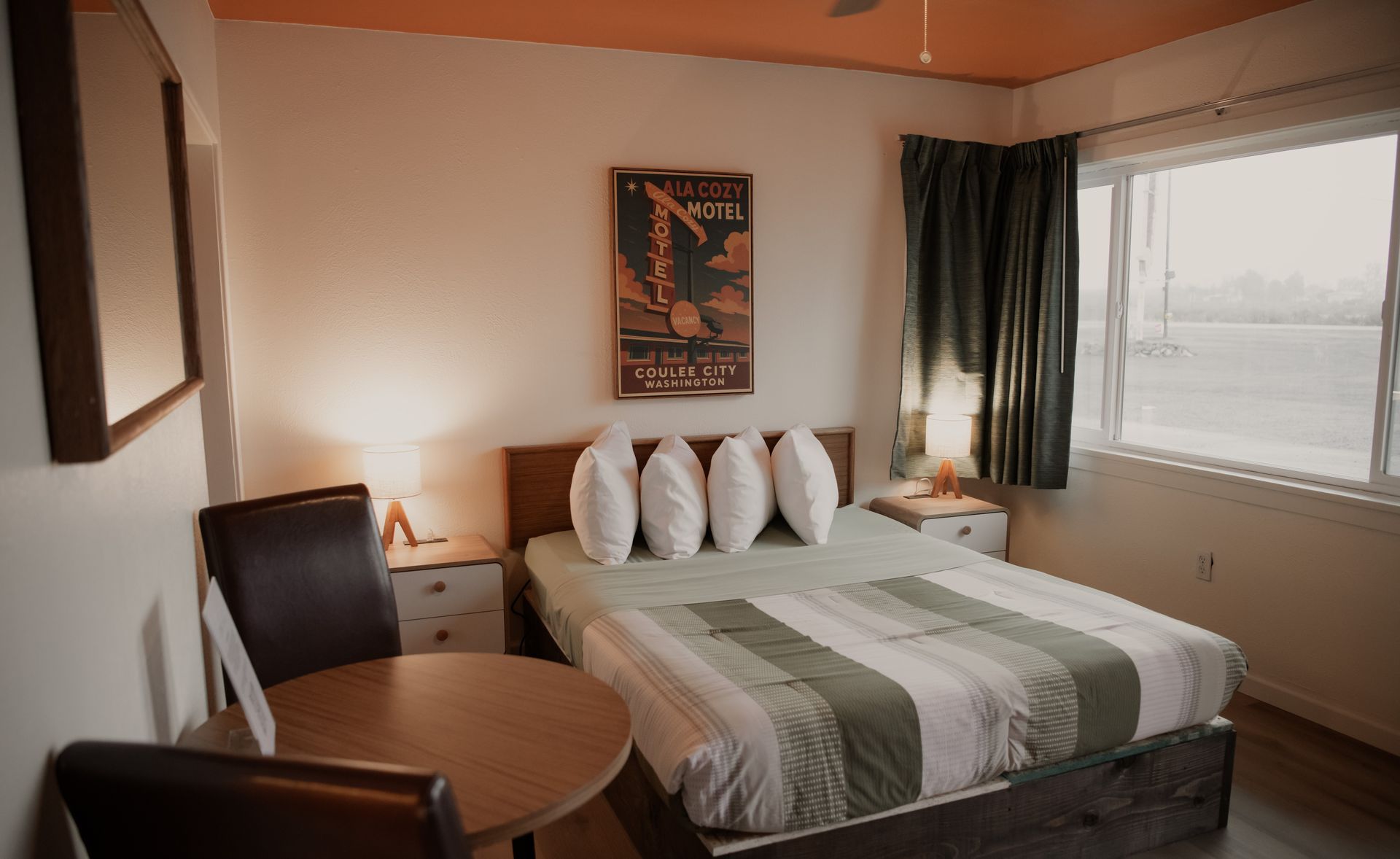A hotel room with a bed, table, and window. The room has neutral tones with a vintage poster.