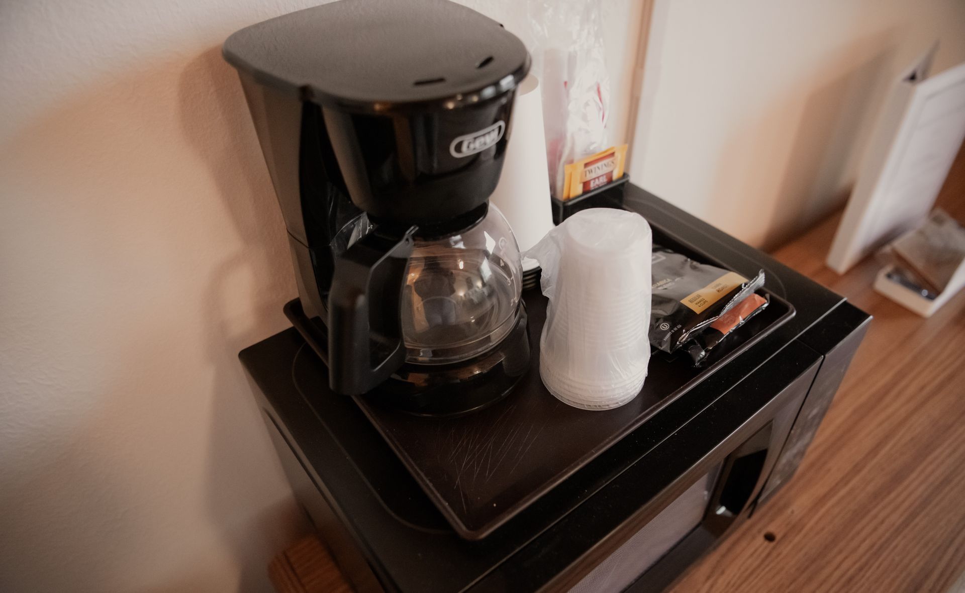 Coffee maker, cups, and condiments on a small microwave in a room, next to a wall.