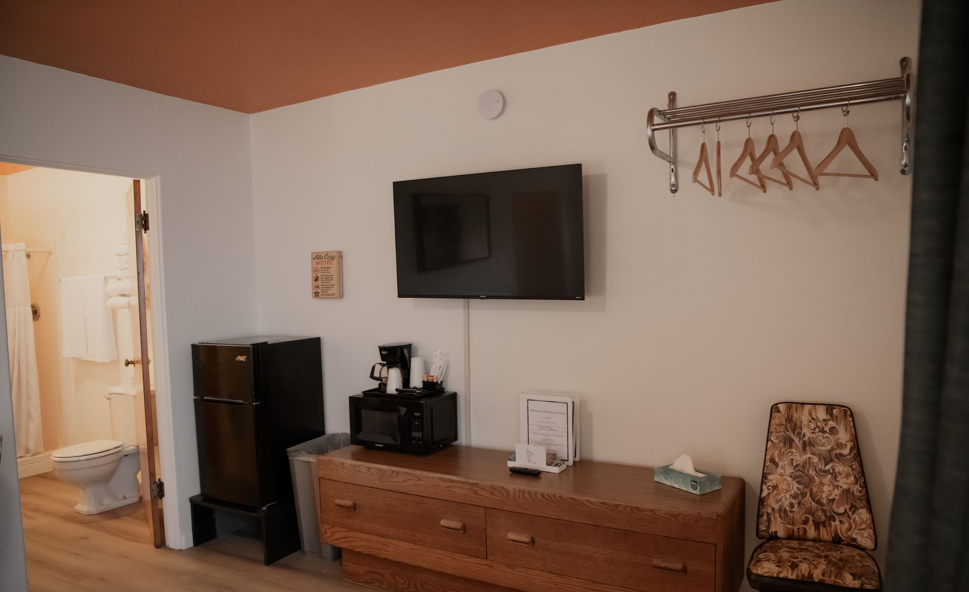Hotel room interior with TV, dresser, mini-fridge, and chair. A doorway leads to a bathroom.