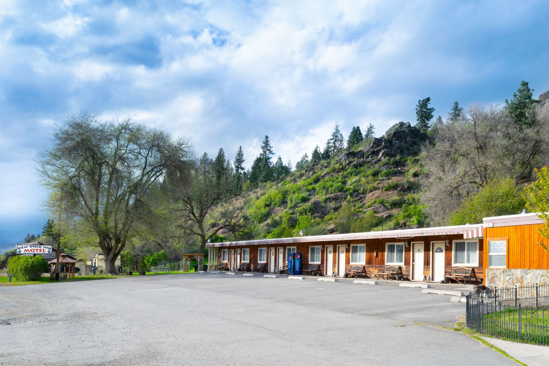 Motel with single-story rooms, wooden facades, and a parking lot, set against a hillside with trees and a cloudy sky.