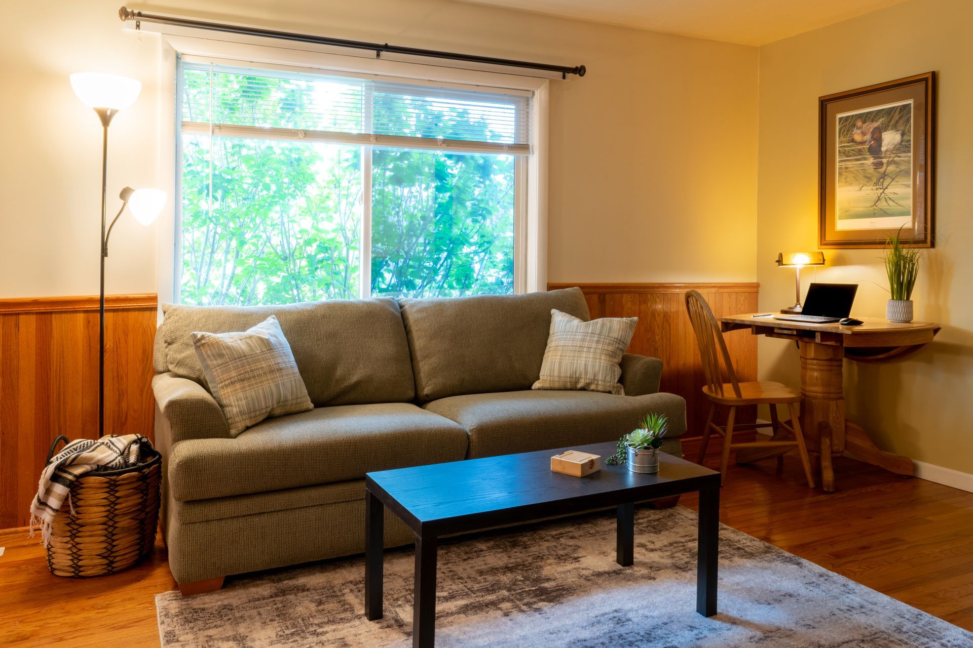 Cozy living room with a green couch, small desk with a laptop, and a large window.