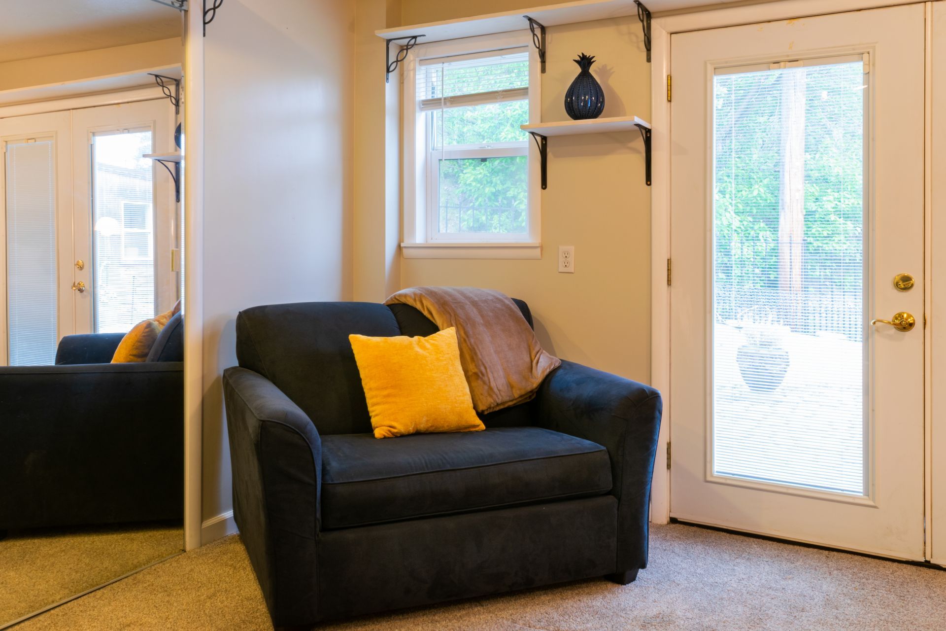 A cozy corner with a navy armchair, a yellow pillow, and a window with a shelf holding a blue vase, near a door.