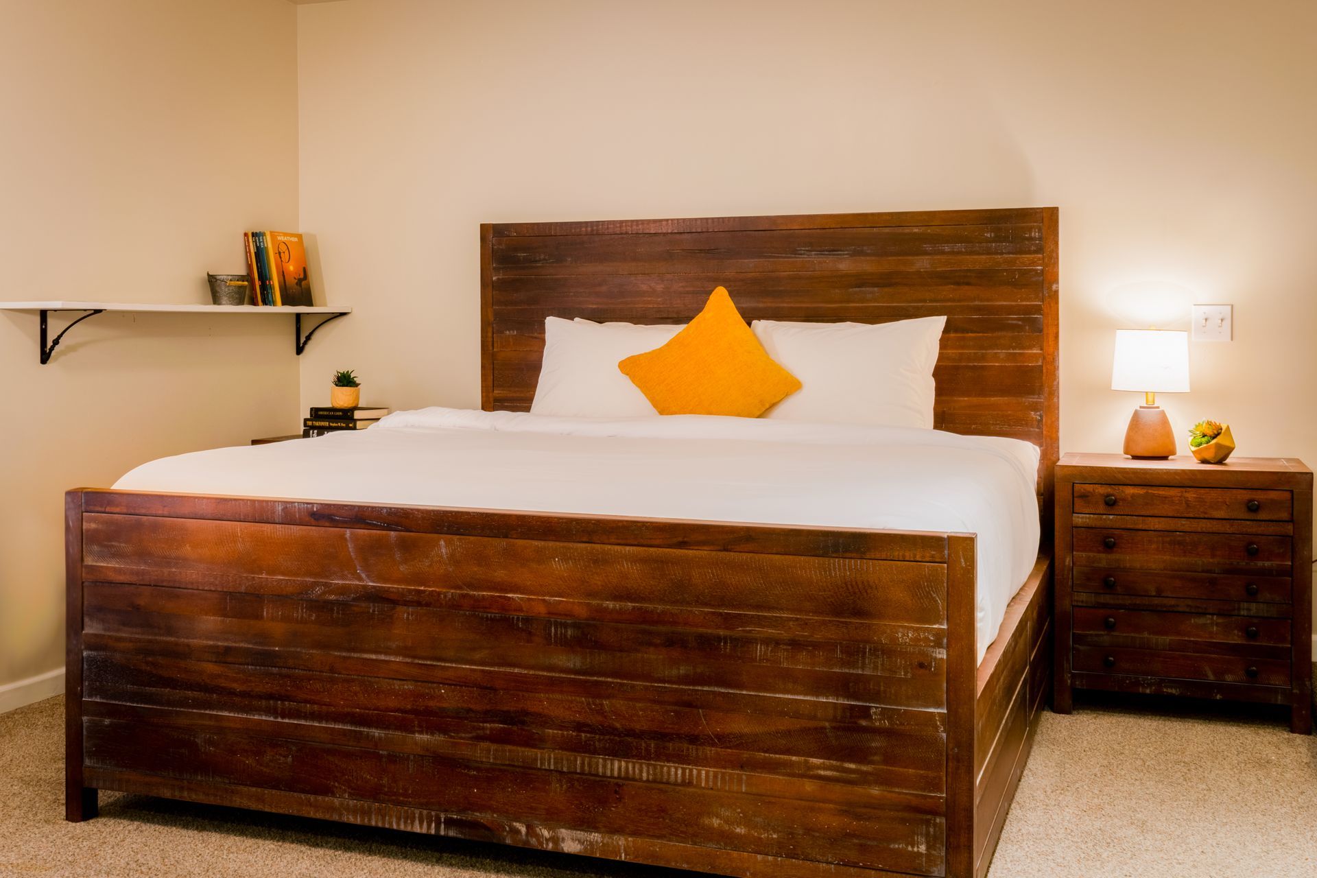 A wooden bed with a white comforter and yellow pillow in a bedroom with a nightstand and shelf.