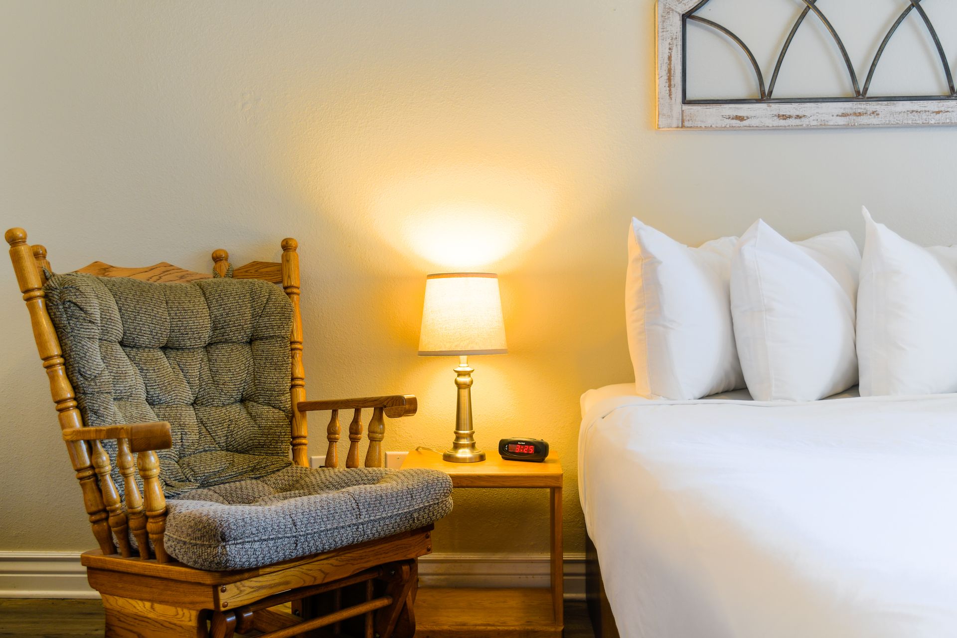 A bedroom with a wooden rocking chair, lamp on a nightstand, and a bed with white linens.