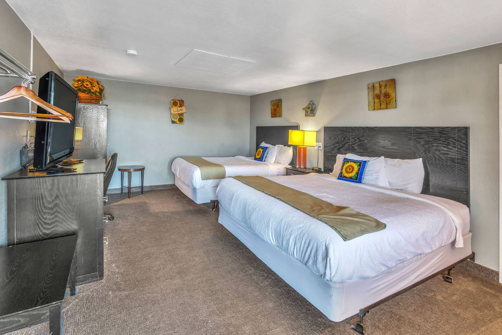 Hotel room with two queen beds, a desk, television, and sunflower decor on walls.