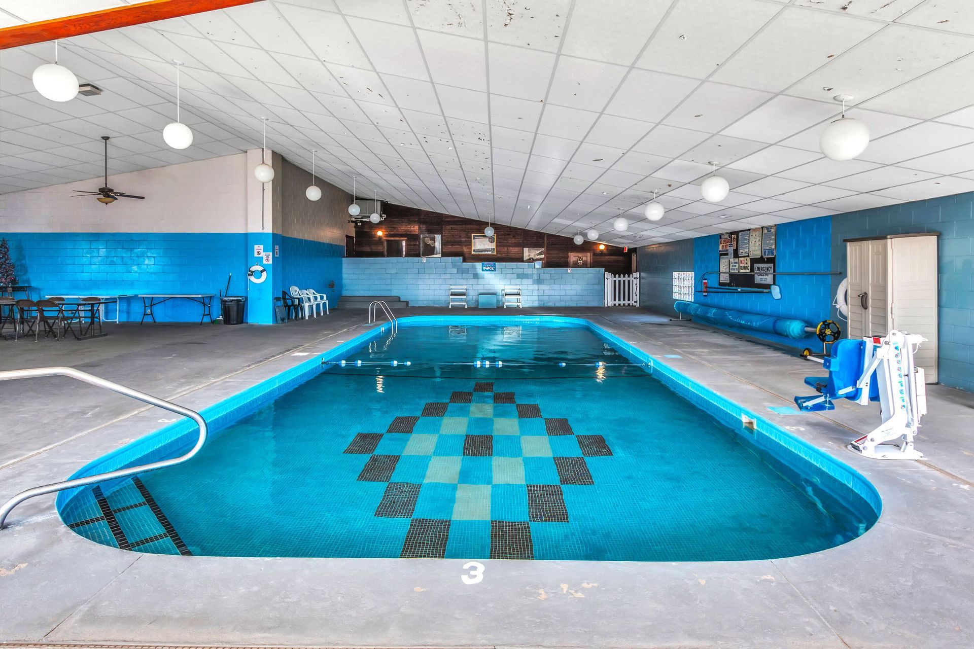 Indoor swimming pool with blue water and checkered pattern, white ceiling, and gray floor.