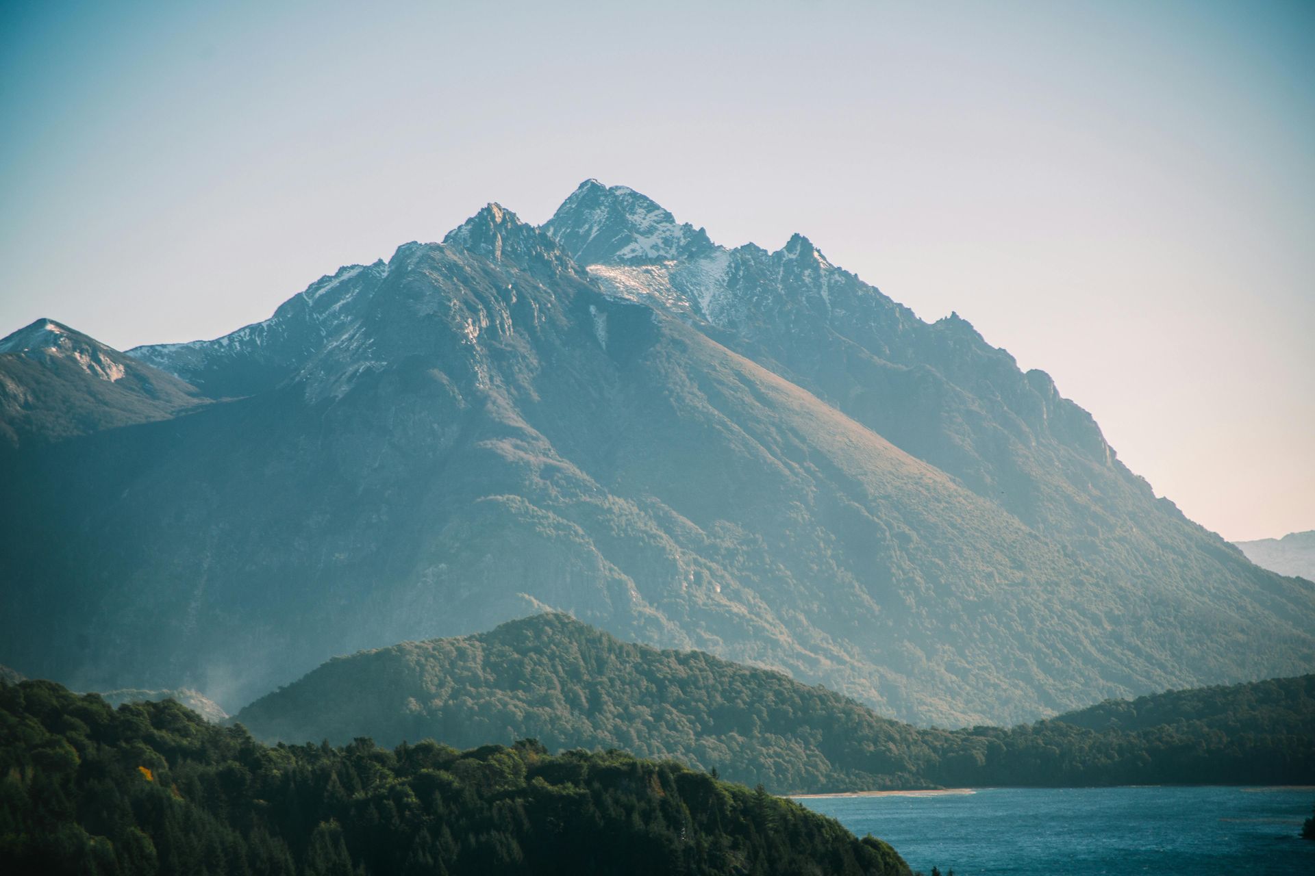 Two mountain goats with curved horns stand on a rocky ledge, gazing towards the camera, against a blue sky with white clouds.