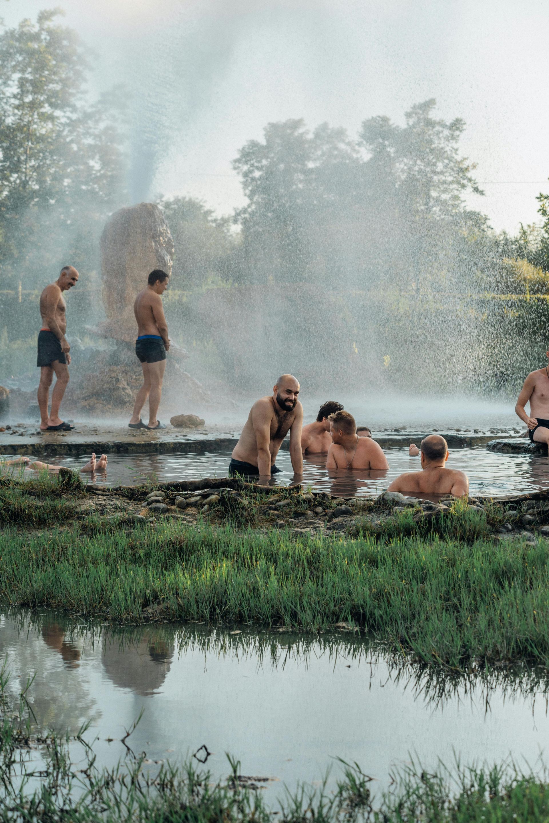 Men in swimwear relaxing in a natural hot spring with steam rising. Green foliage surrounds the water.