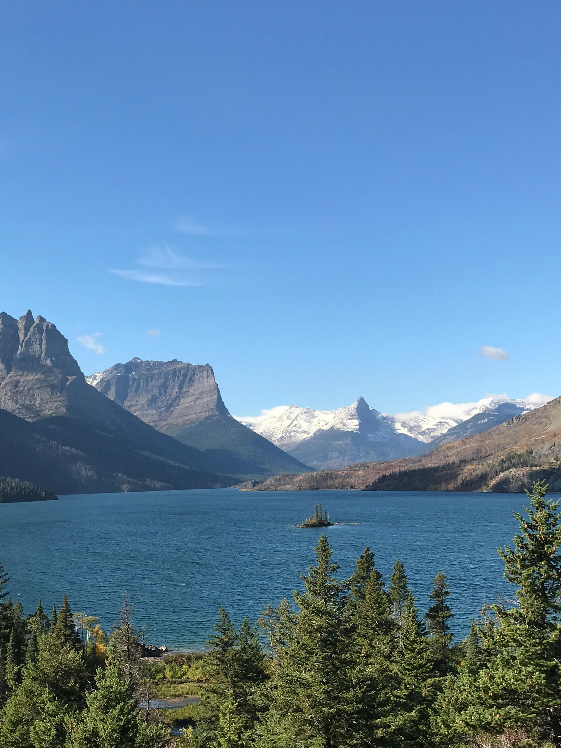 Calm lake scene with people in a boat, a small hut, and mountains reflected in the water under a misty sky.