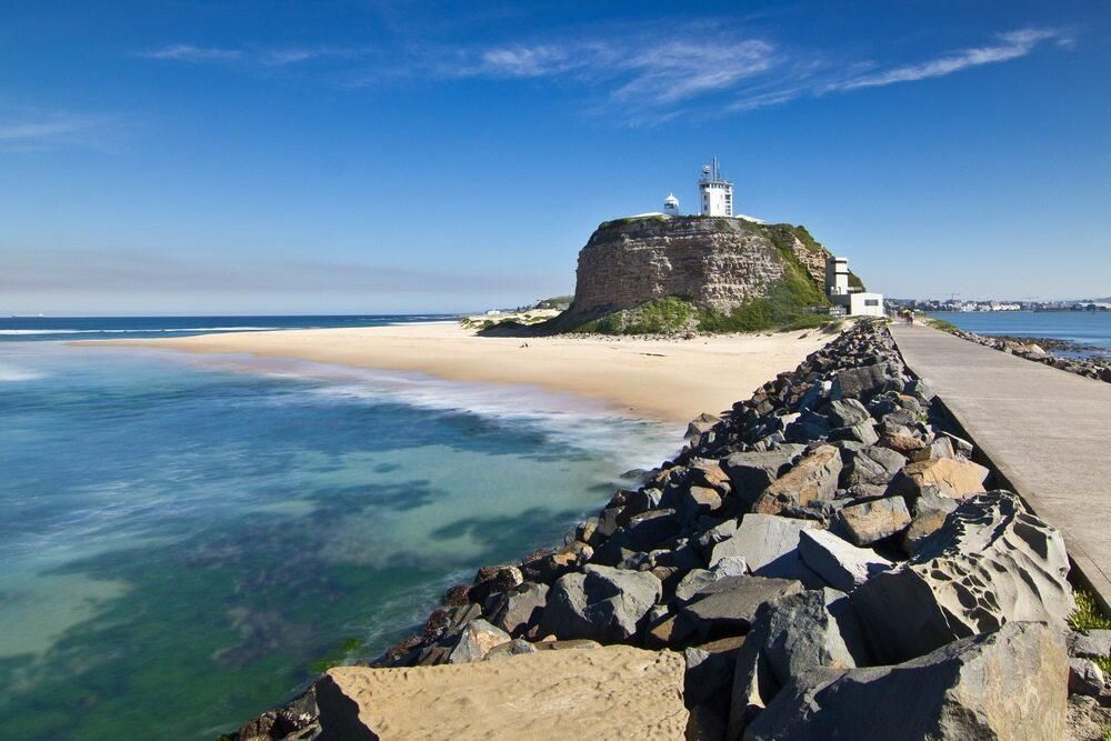 A Pier Leading to a Beach With a Lighthouse on Top of a Rocky Hill — Kymar Steel Fabrications in Newcastle, NSW